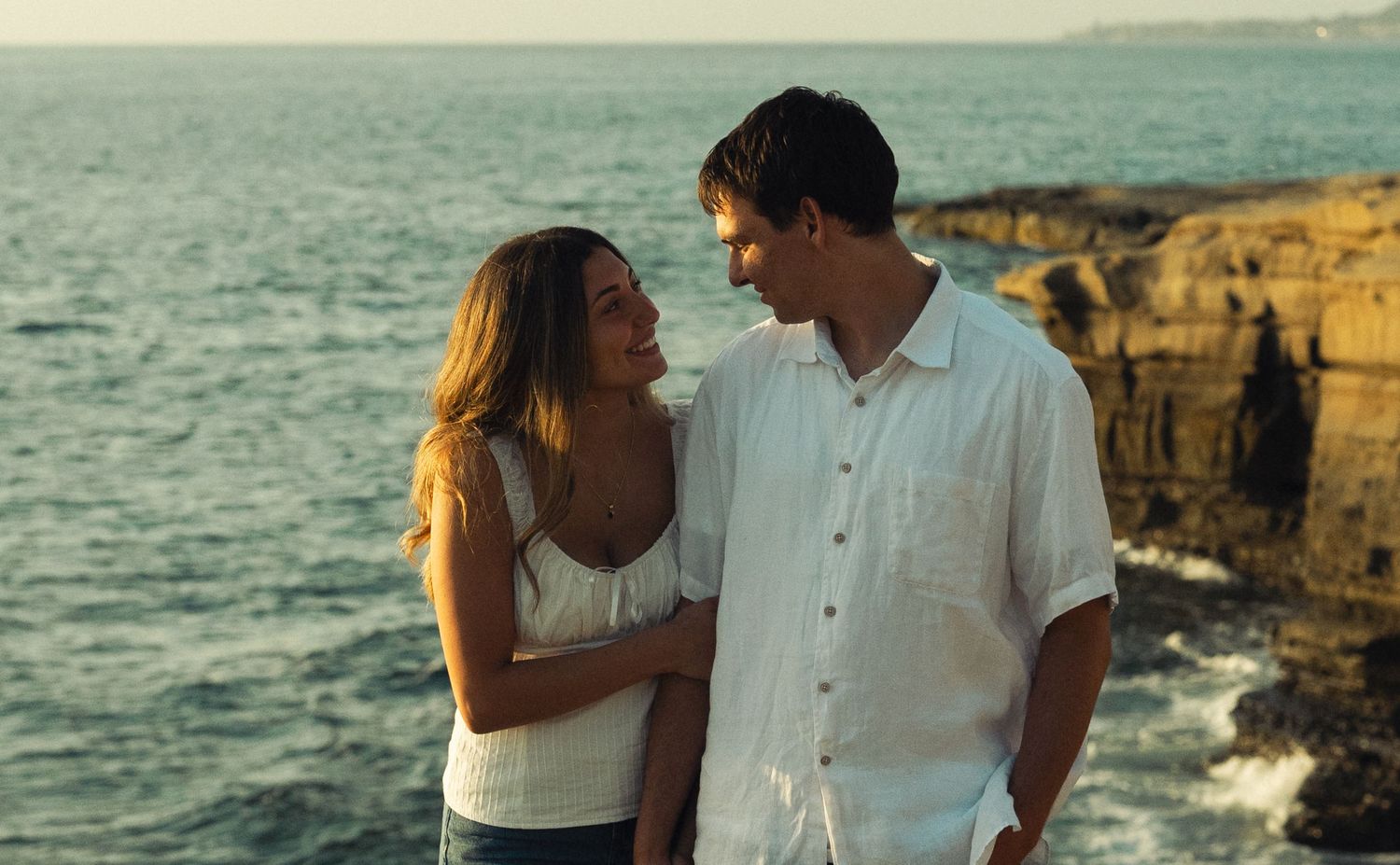 Romantic couple smiling at each other on rocky coastal cliffs at sunset near the ocean.