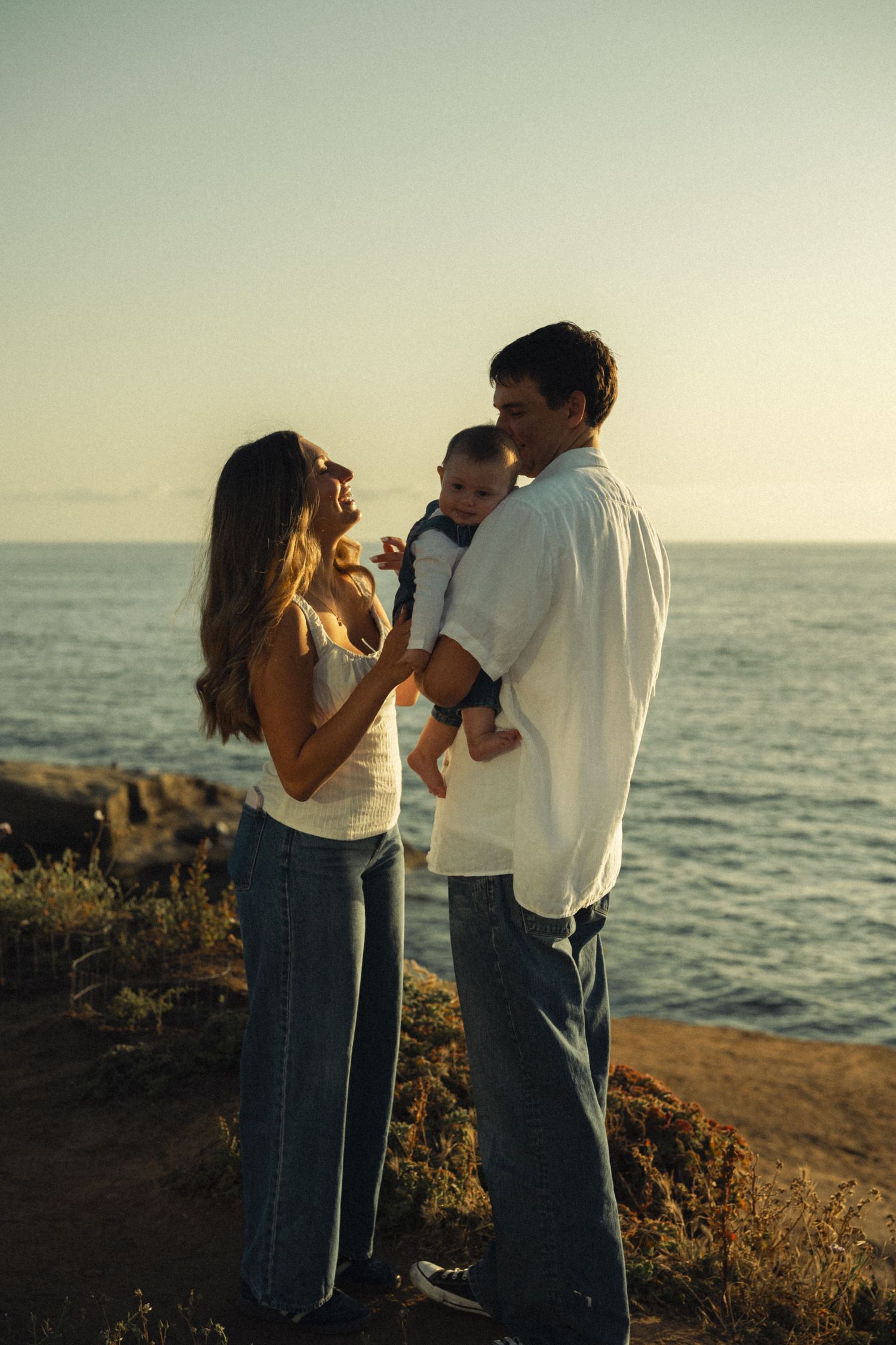 Happy young family with baby enjoying a beautiful ocean sunset on rocky cliffs.