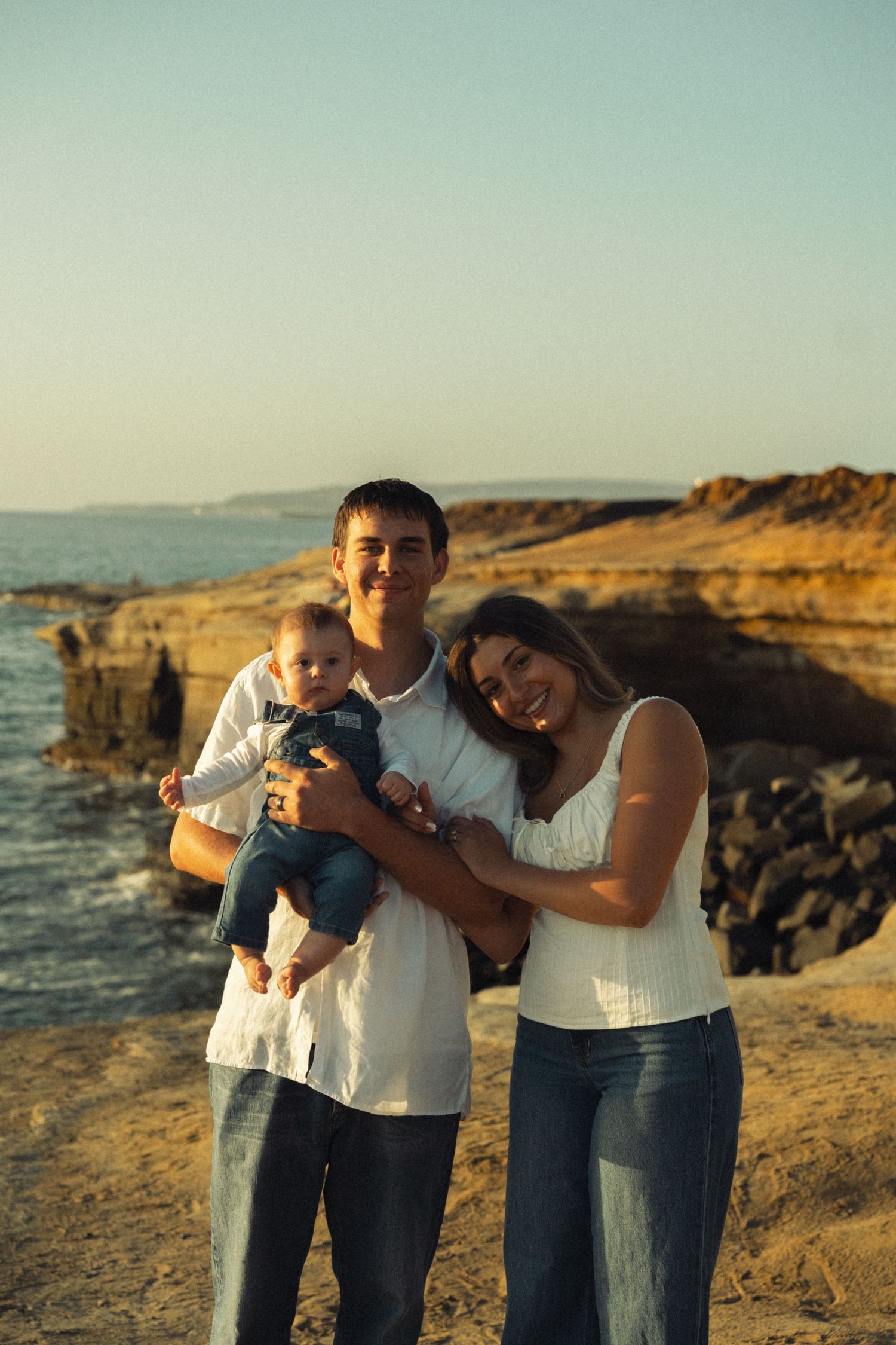 Young family with baby posing on rocky coastal shore at golden hour, parents in white tops and jeans smiling warmly.
