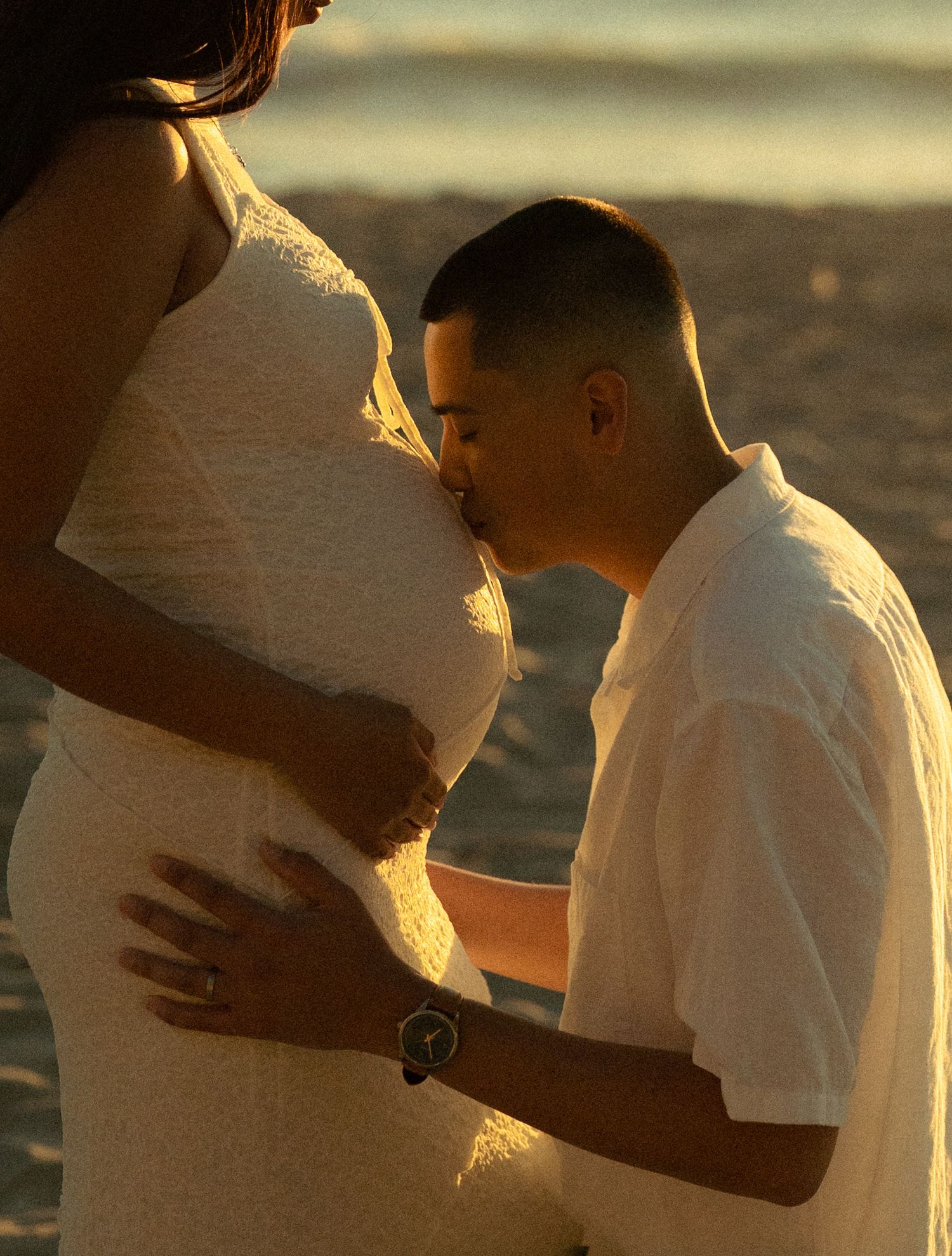 Man kissing pregnant woman's belly on a beach at golden hour, both wearing white outfits.