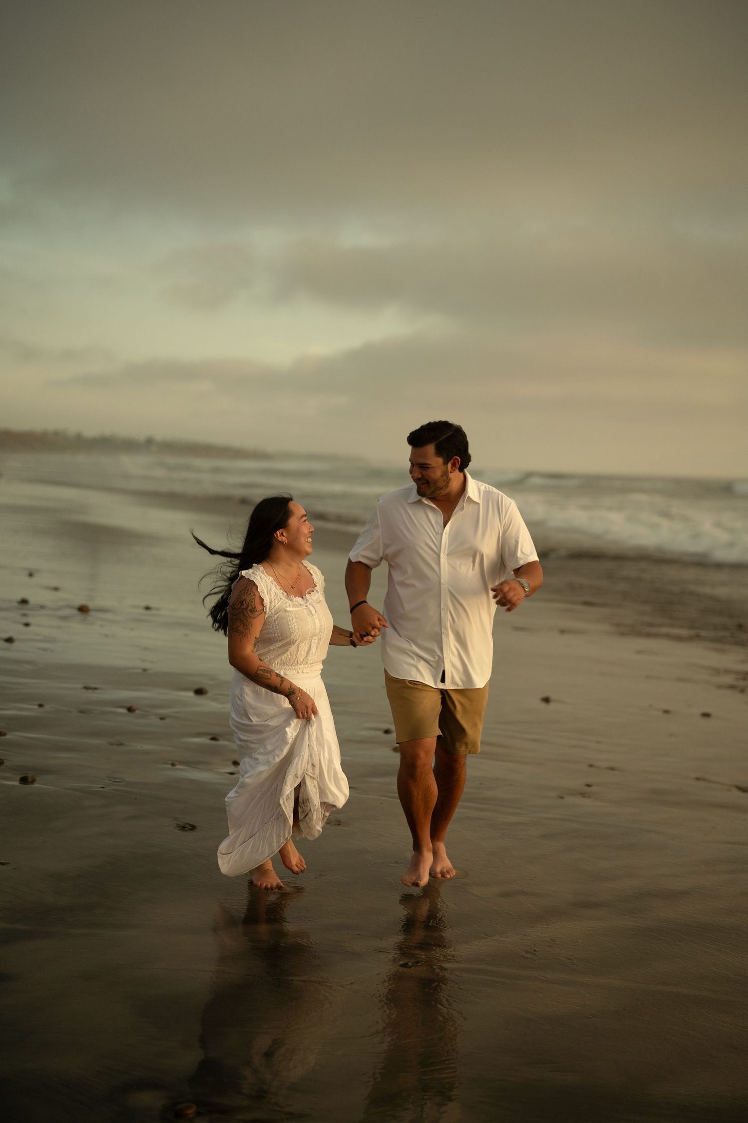 Happy couple running barefoot hand-in-hand along wet sandy beach at dusk, woman in white dress, waves behind them.