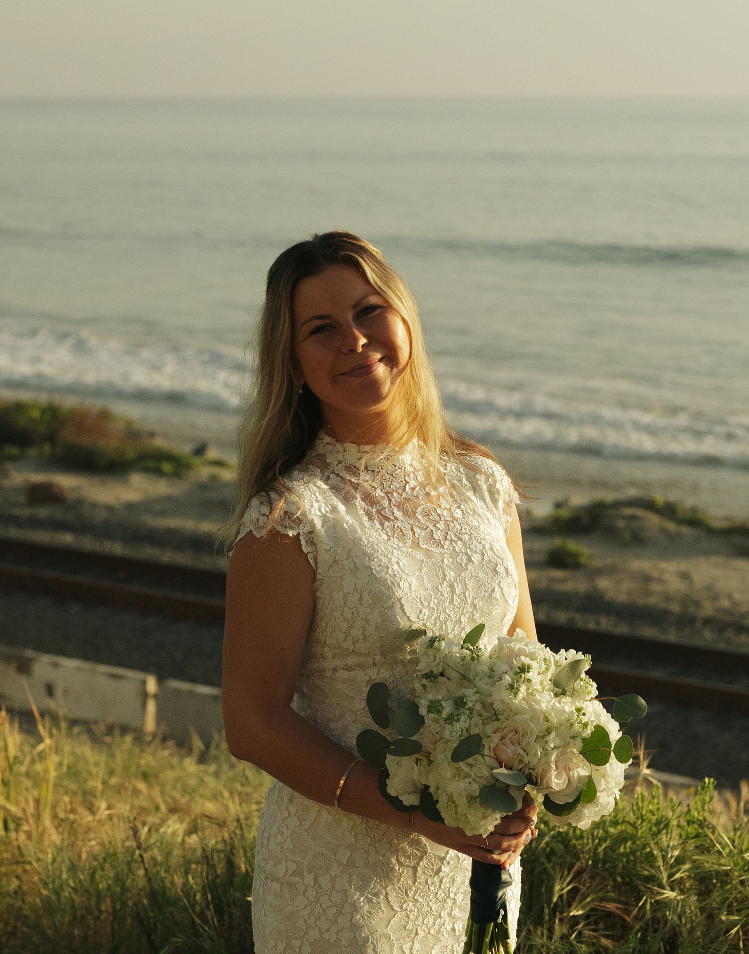 Blonde bride in lace high-neck wedding gown holds white floral bouquet smiling at beachside sunset.