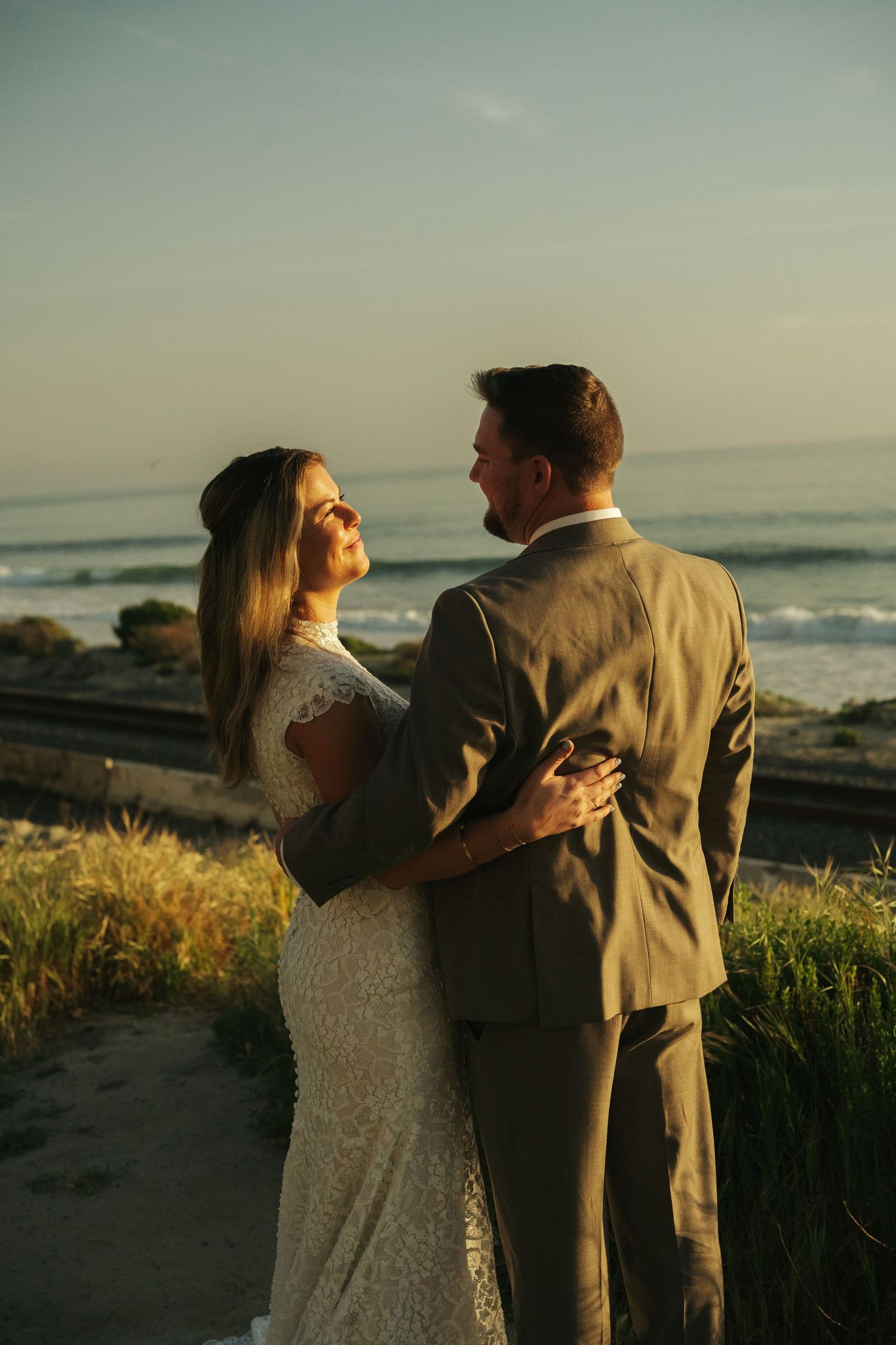 Newlyweds embracing at sunset on a coastal bluff overlooking ocean waves, bride in lace gown smiling at groom.