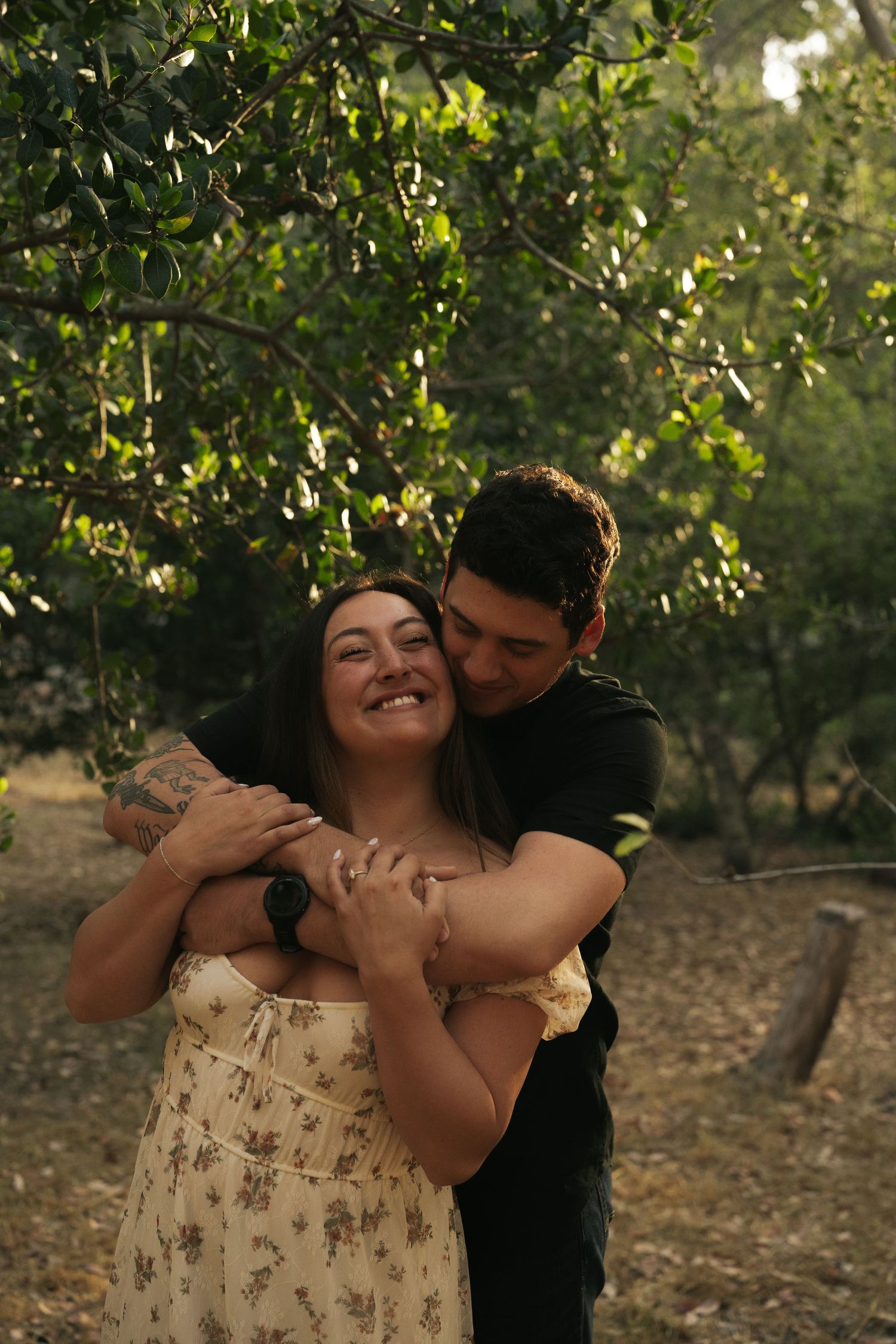 Man in black shirt embracing woman in floral dress from behind under green trees outdoors.