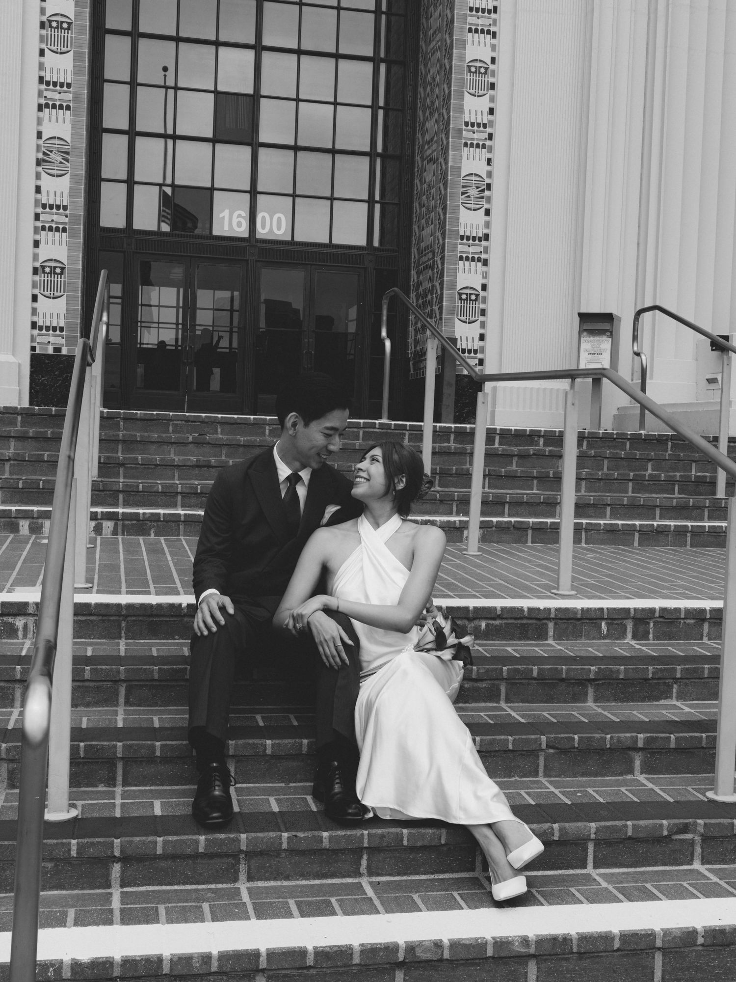 Black and white photo of a couple in wedding attire sitting on brick steps outside a building, sharing a romantic moment.