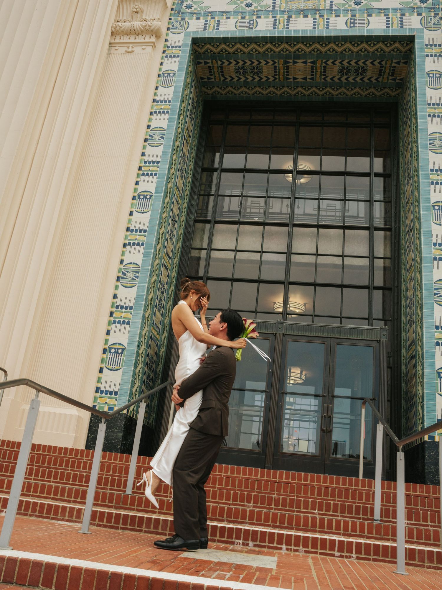 Groom lifting bride in white dress on brick steps before ornate tiled building entrance during wedding photo session.
