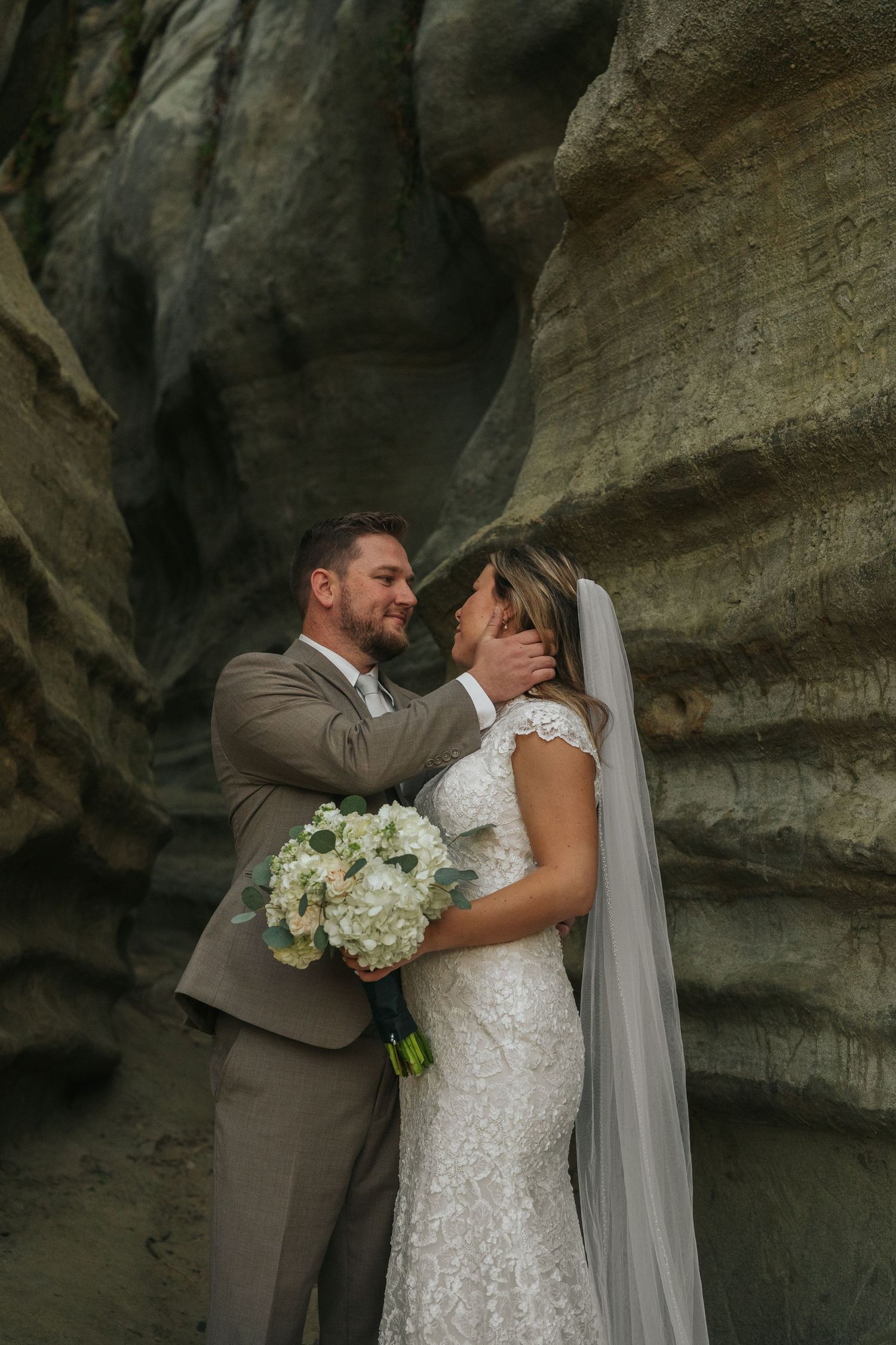 Groom tenderly holds brides face in romantic sandstone canyon wedding portrait with white floral bouquet.