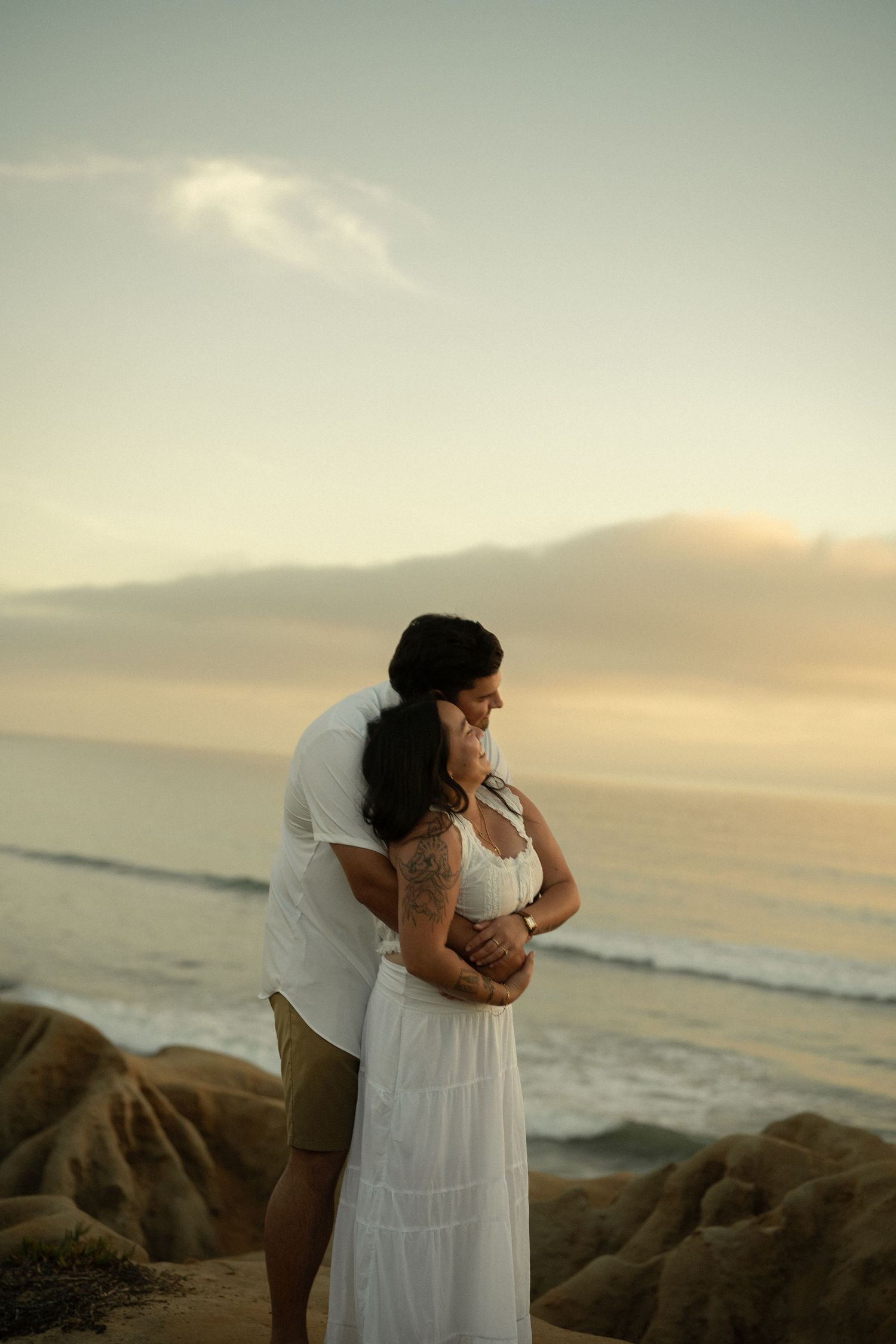 Man hugging woman from behind on rocky beach at sunset, both gazing at calm ocean waves in warm evening light.