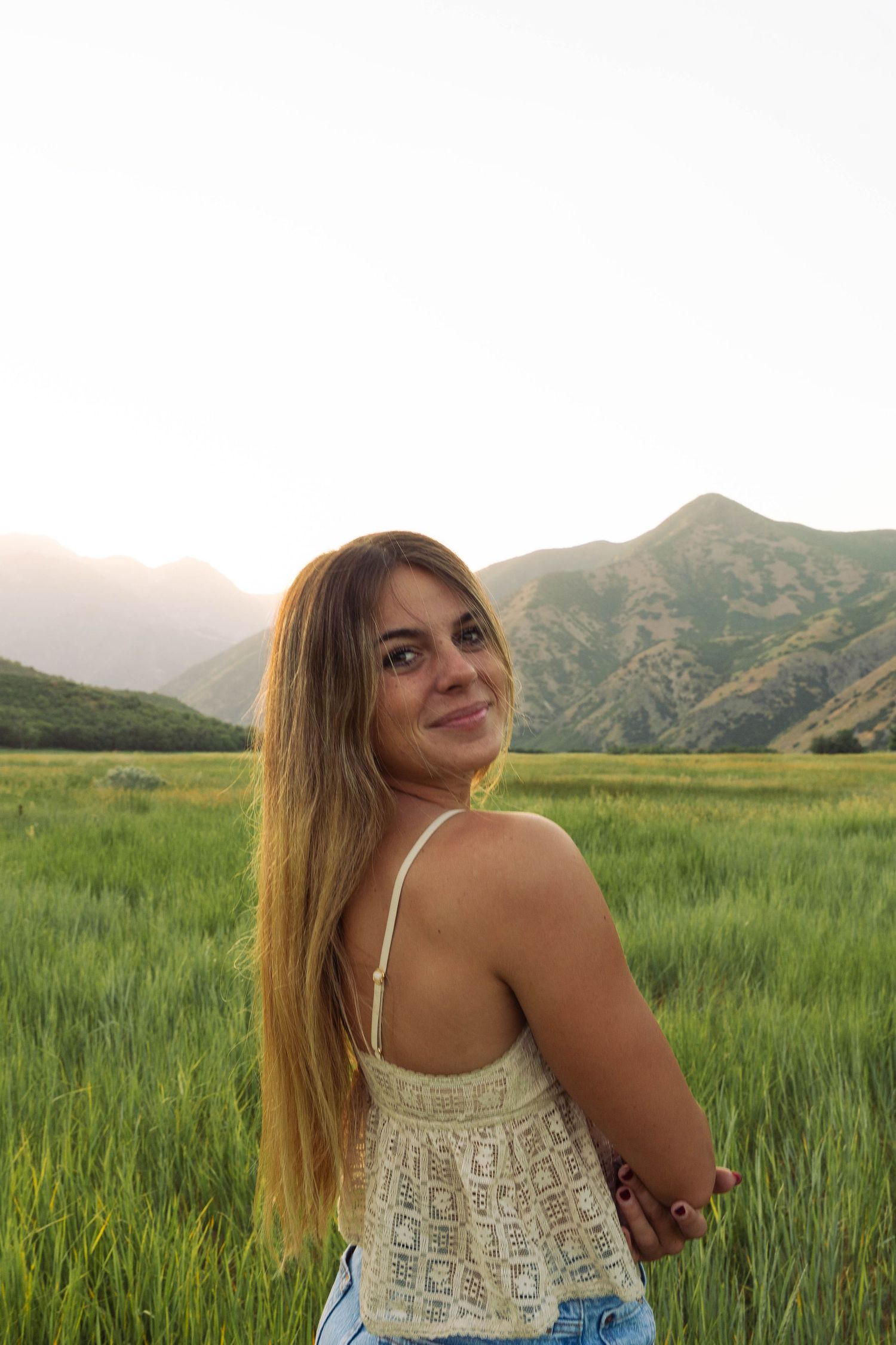 Young woman with long hair smiling over her shoulder in a green field with mountains at sunset.