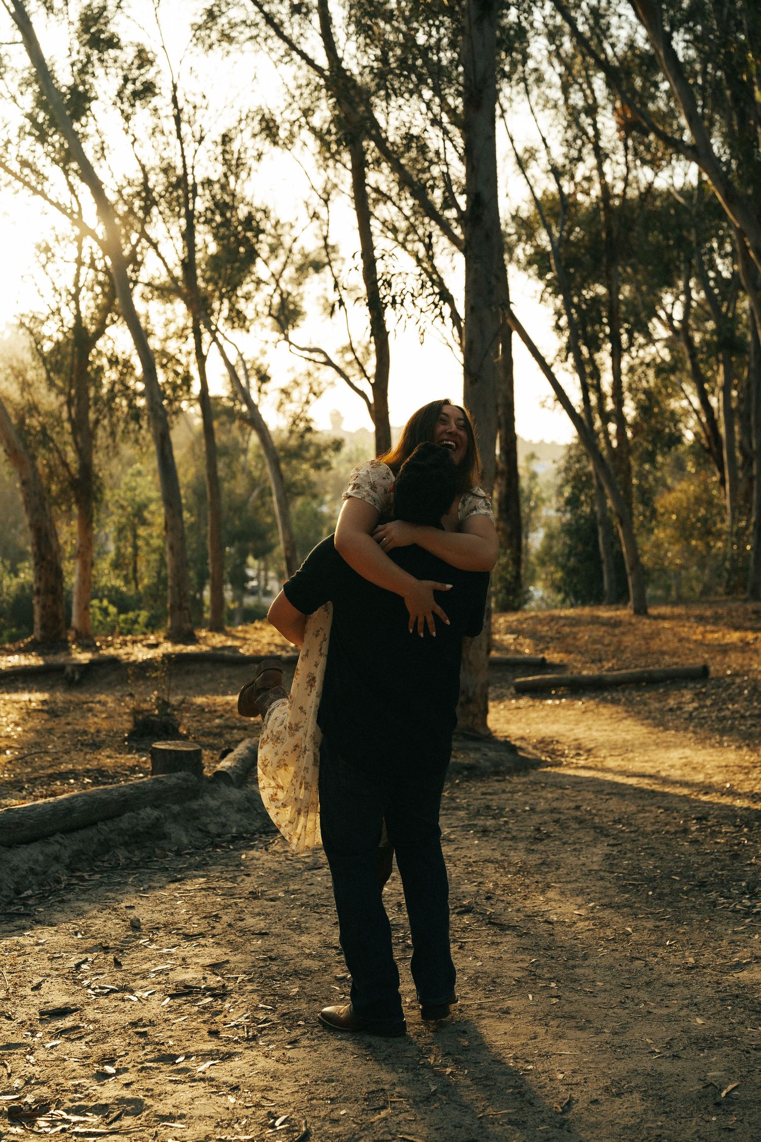 A man lifts and embraces a woman in a floral dress in a golden sunlit park setting.