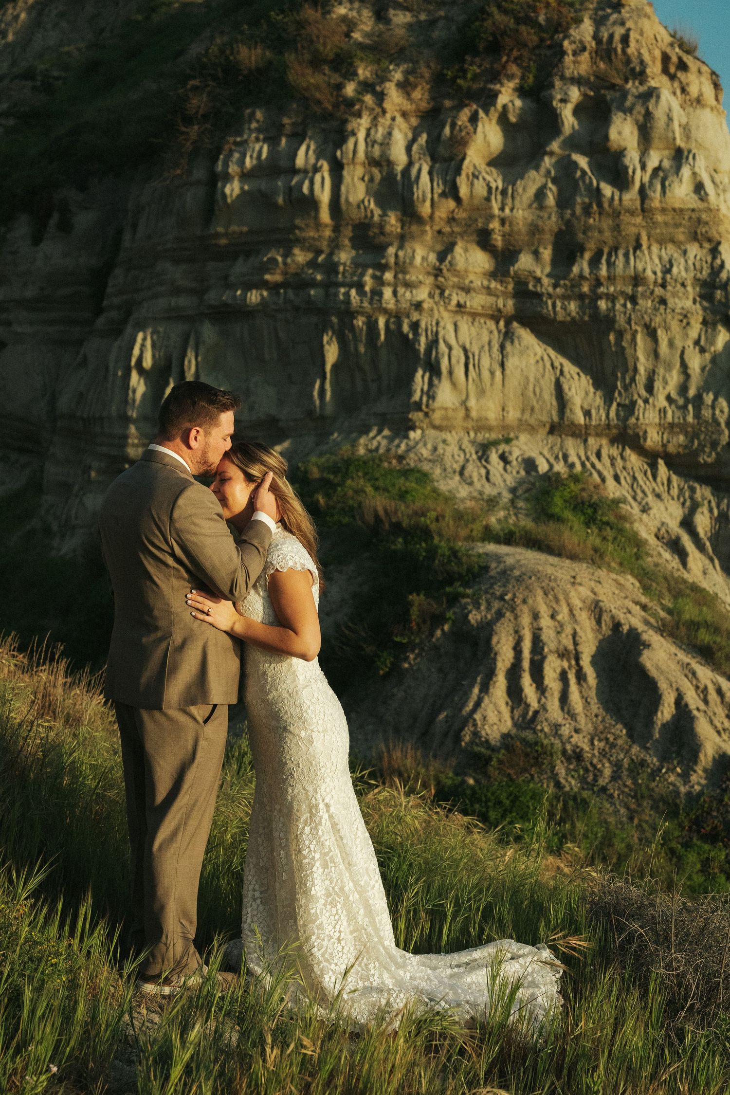 Wedding couple embraces tenderly in front of dramatic eroded rock cliffs in warm golden evening light.