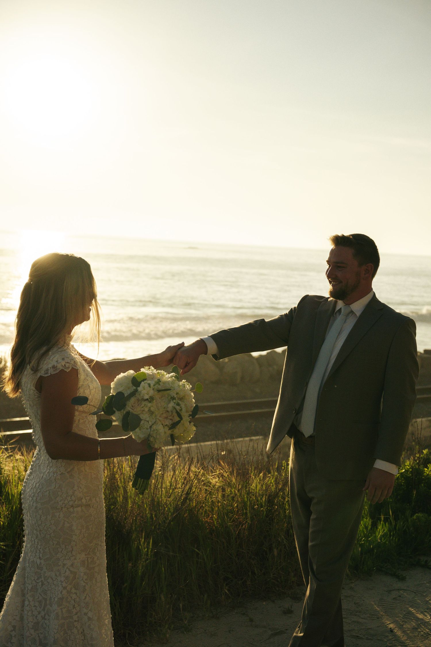 Bride and groom holding hands at sunset near the ocean, bride holding white bouquet in a lace dress.