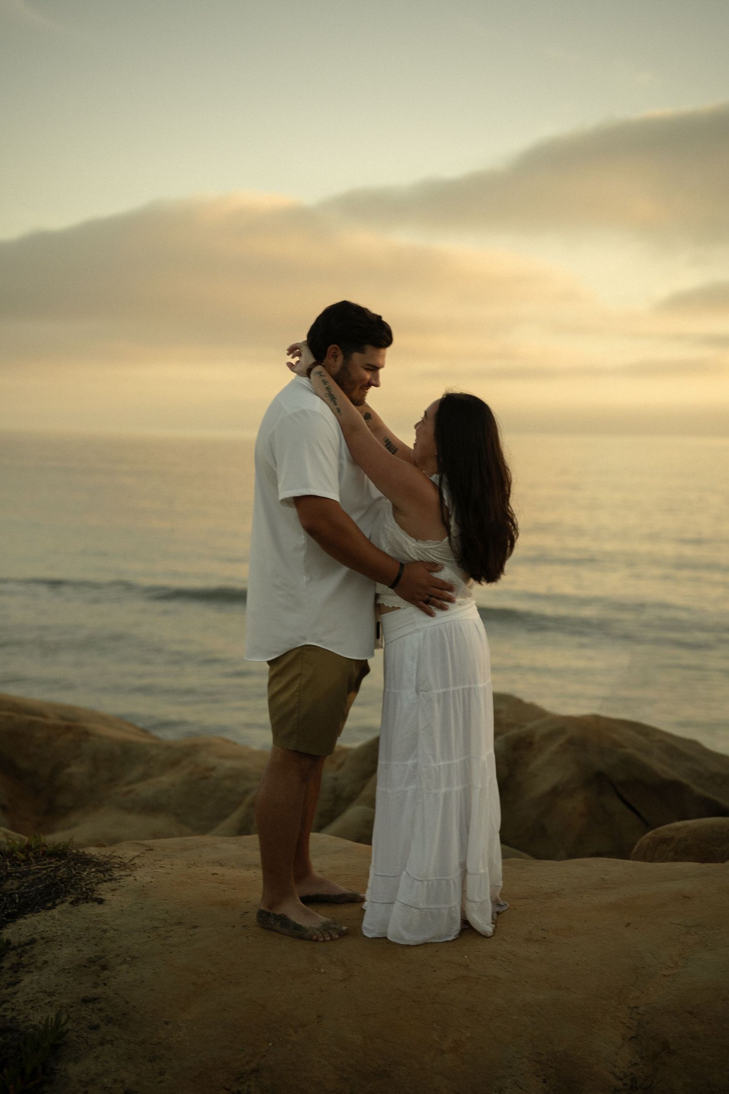 Couple embracing romantically on coastal rocks at golden sunset, woman in white dress touching man's face by the ocean.