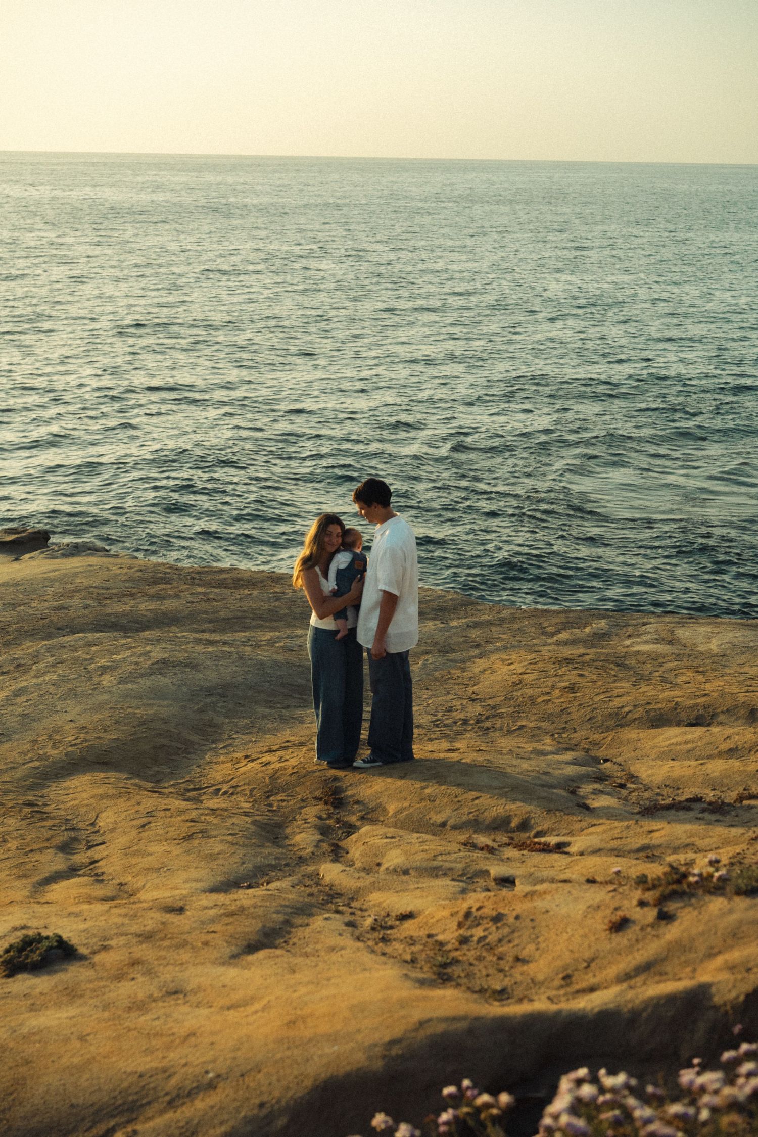 Young couple holding baby standing on sandstone cliffs overlooking a calm ocean at dusk.