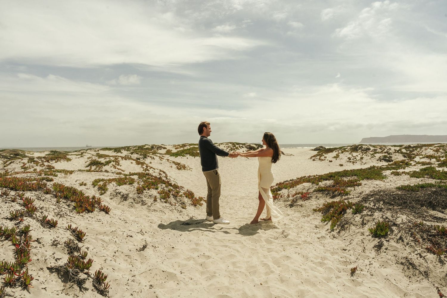 A couple holds hands and dances together on a sandy beach with dunes and greenery under a cloudy sky.