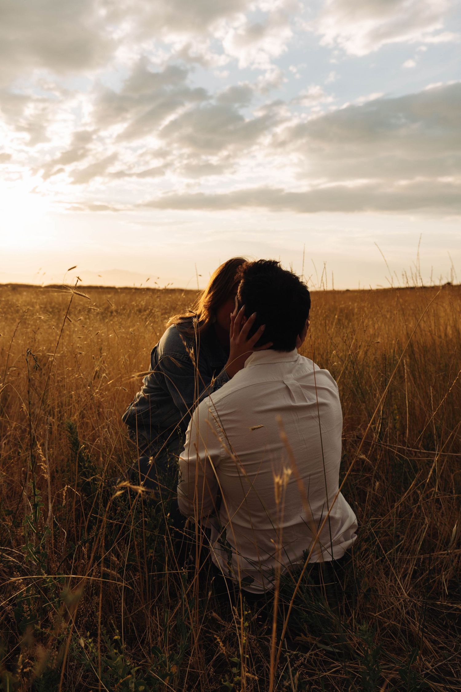 A couple sits close together in a golden wheat field at sunset with dramatic clouds overhead.