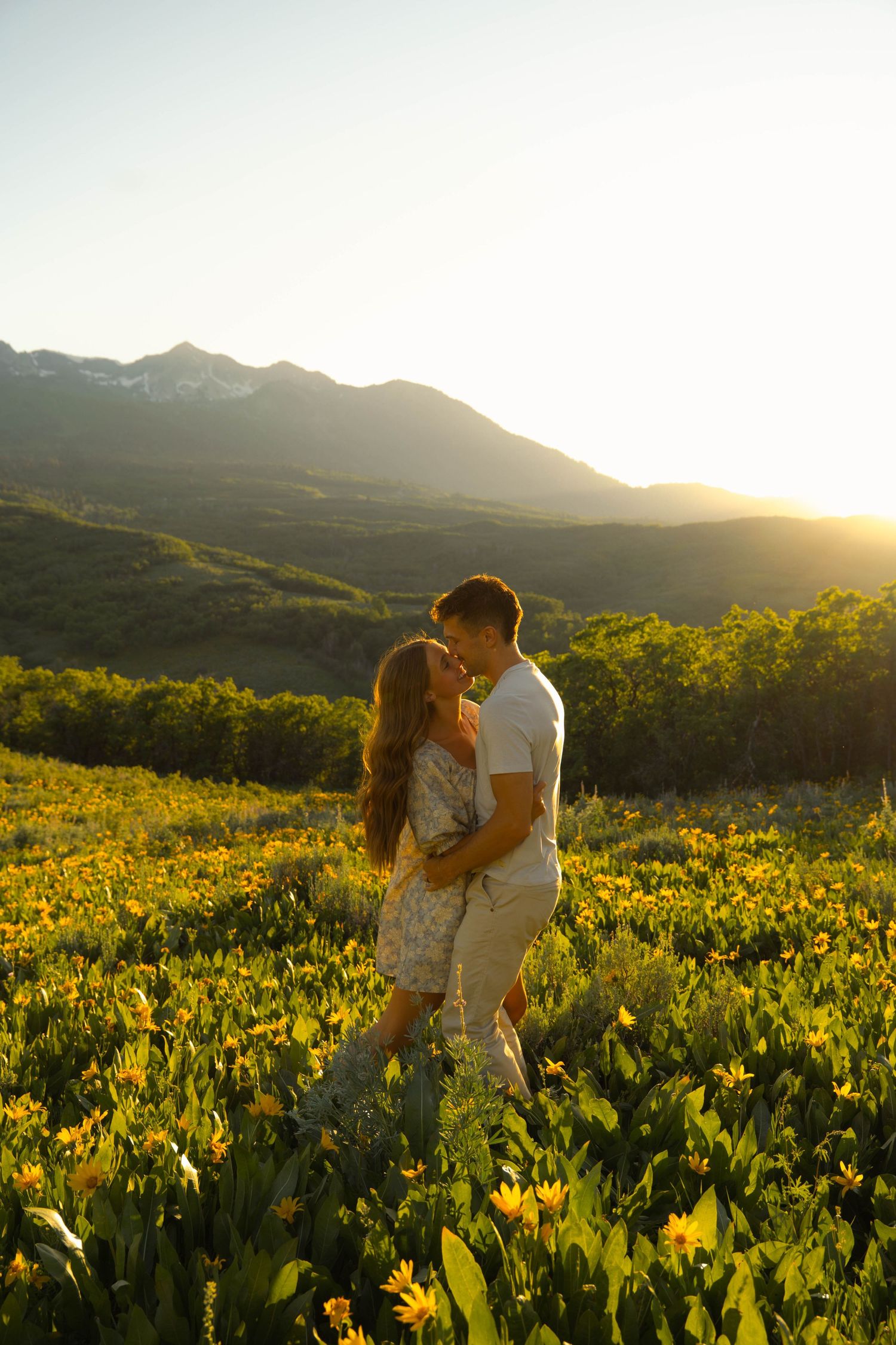 A couple embraces in a field of yellow wildflowers at sunset with mountains in the background.