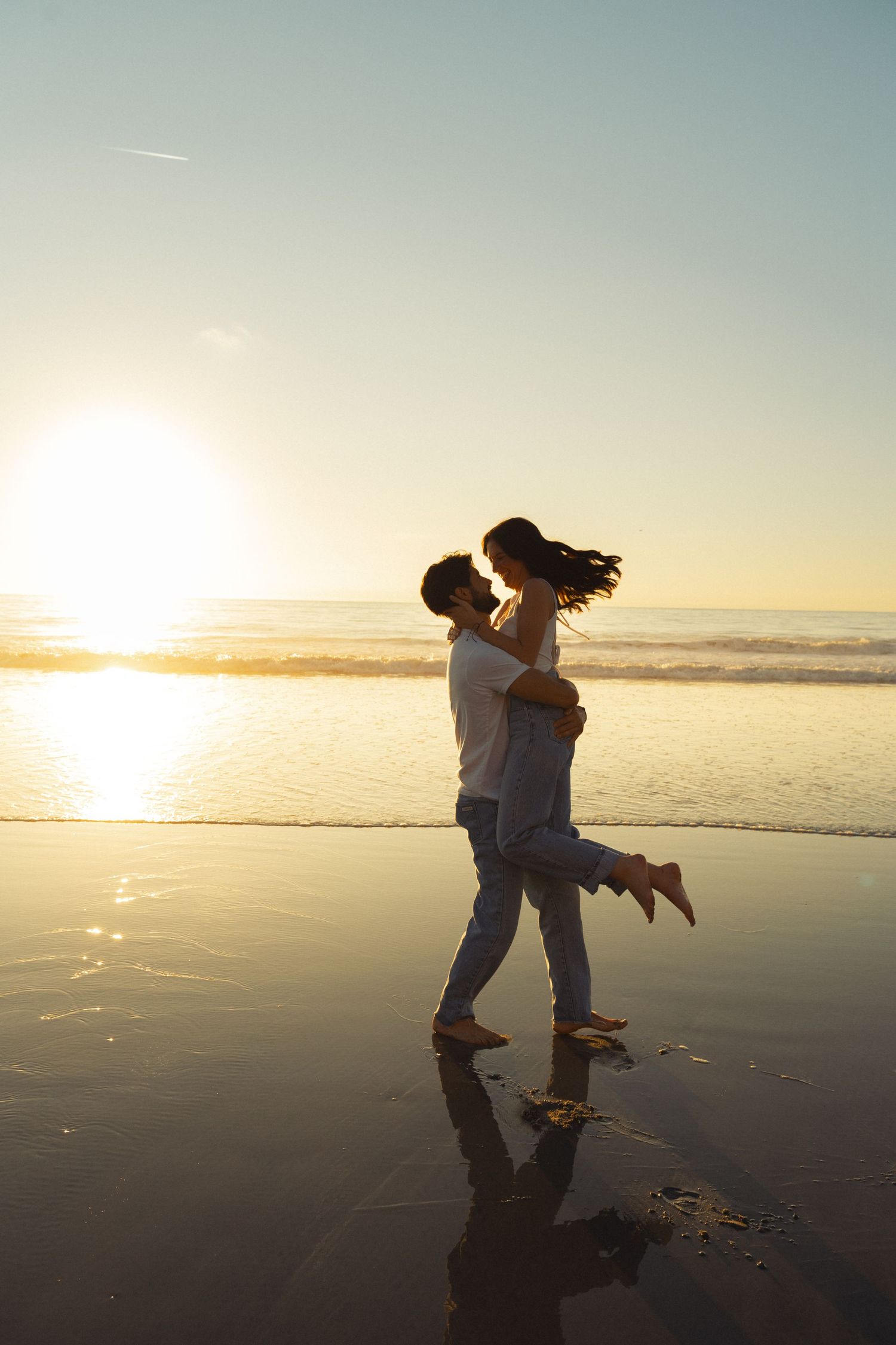 Romantic couple embracing on a sunlit beach at sunset, with golden reflections on wet sand and ocean waves in the background.