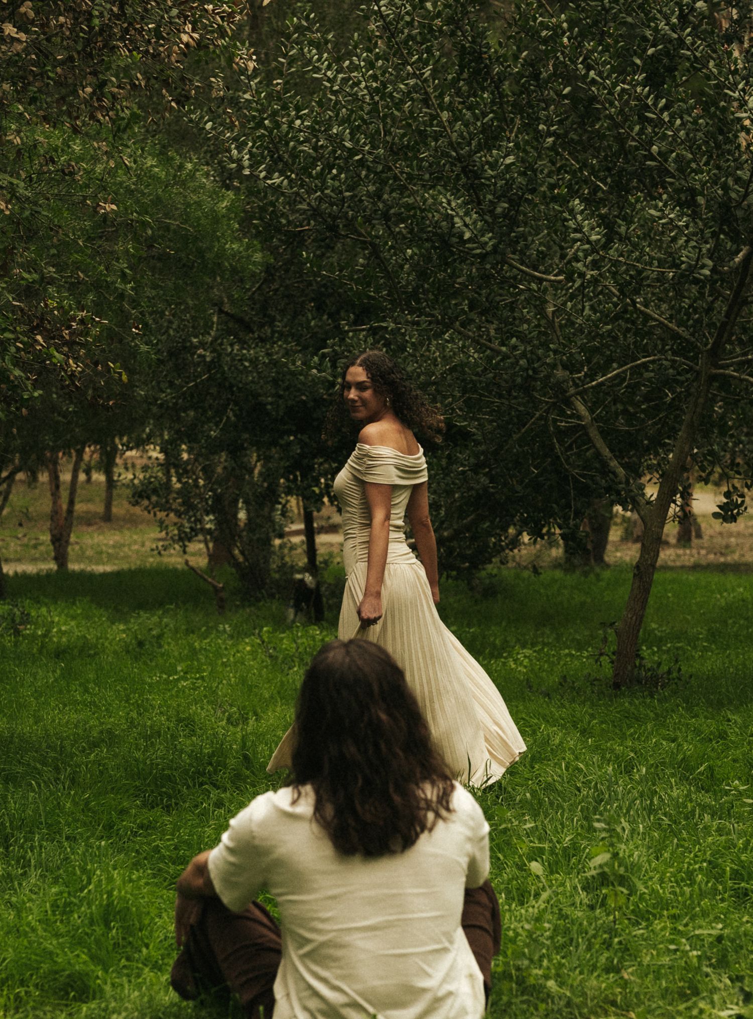 A woman in a flowing white off-shoulder gown stands among green trees as a long-haired person sits in the foreground.