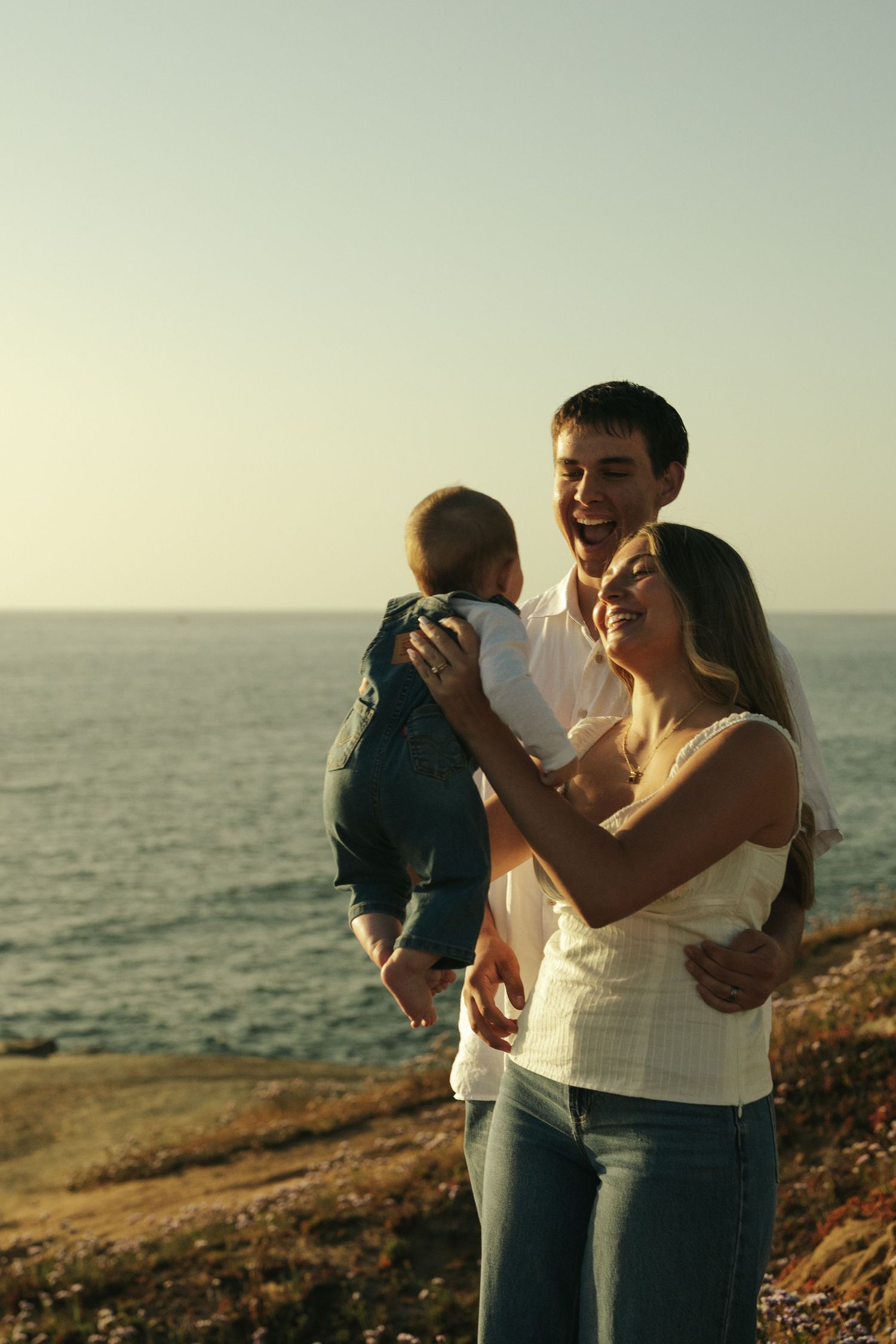Happy young family with baby enjoying a sunset by the ocean, parents smiling and holding their infant child.