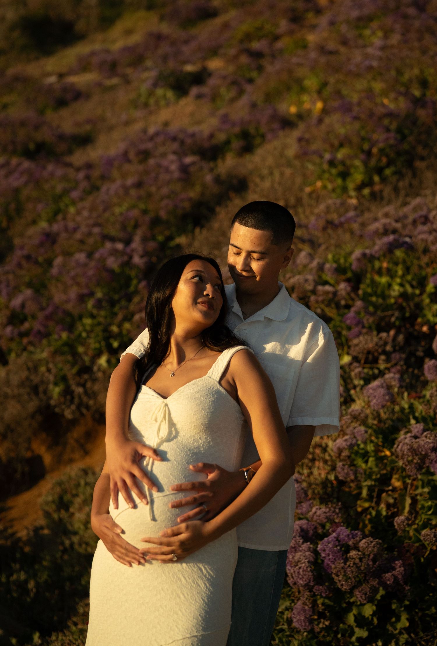 Expectant couple embracing in purple wildflower field at golden hour, man holding pregnant womans belly.