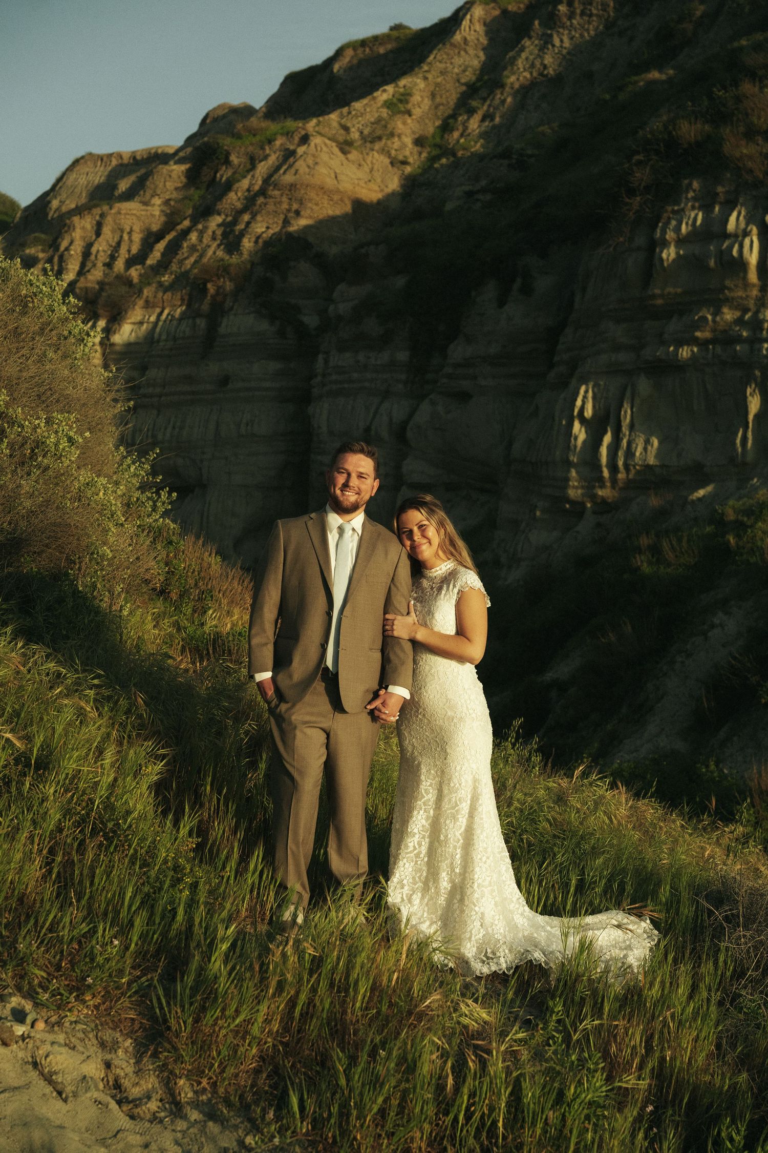 Smiling bride and groom pose together on a sunlit hillside with towering layered rock formations behind them.
