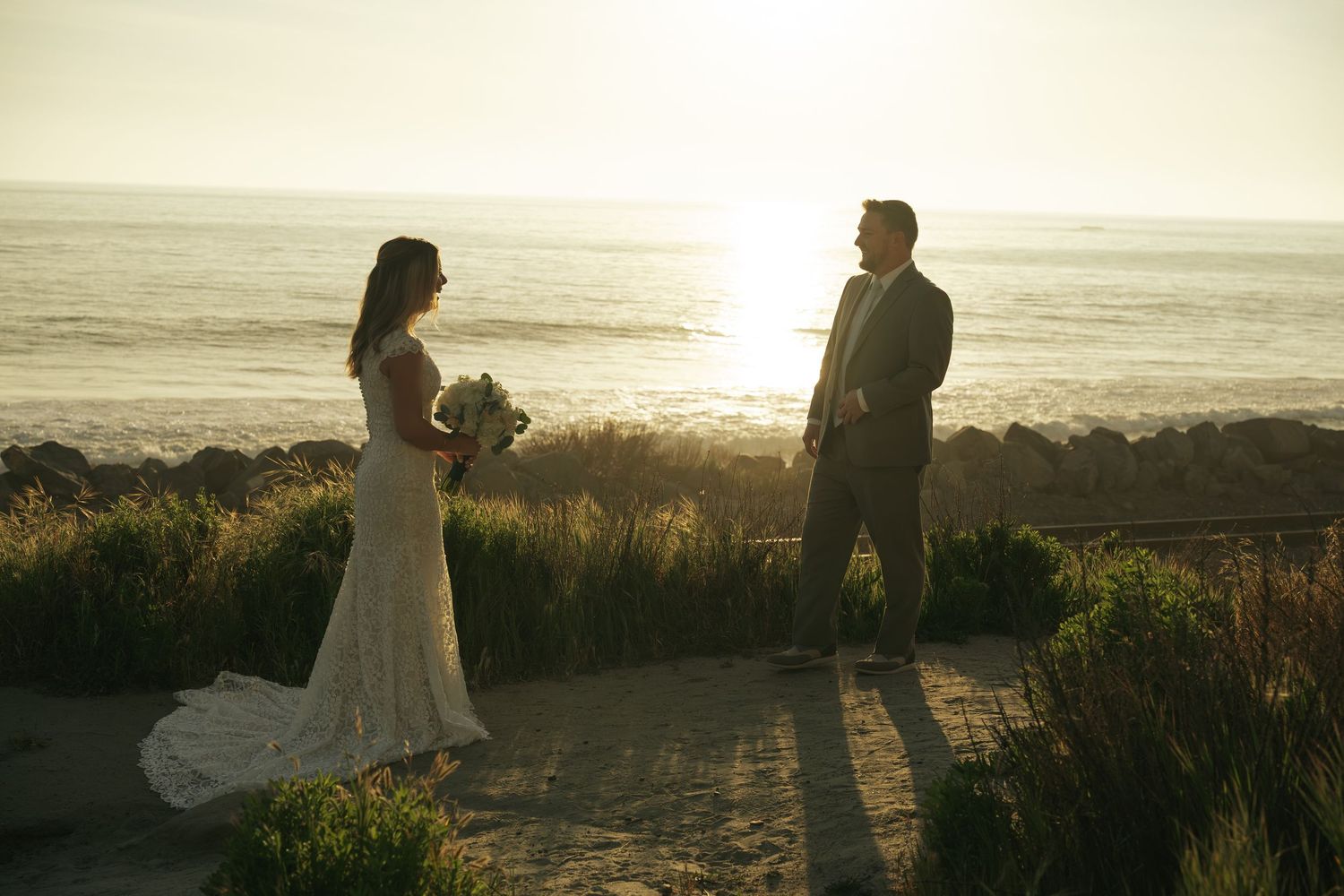 Bride and groom share a romantic sunset moment on a coastal bluff, with ocean waves glowing in the golden light.