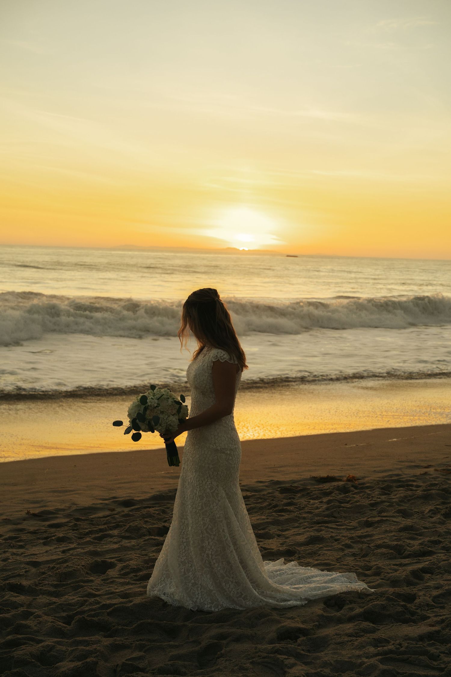 Bride in lace gown holds white bouquet while standing on beach during stunning golden sunset.