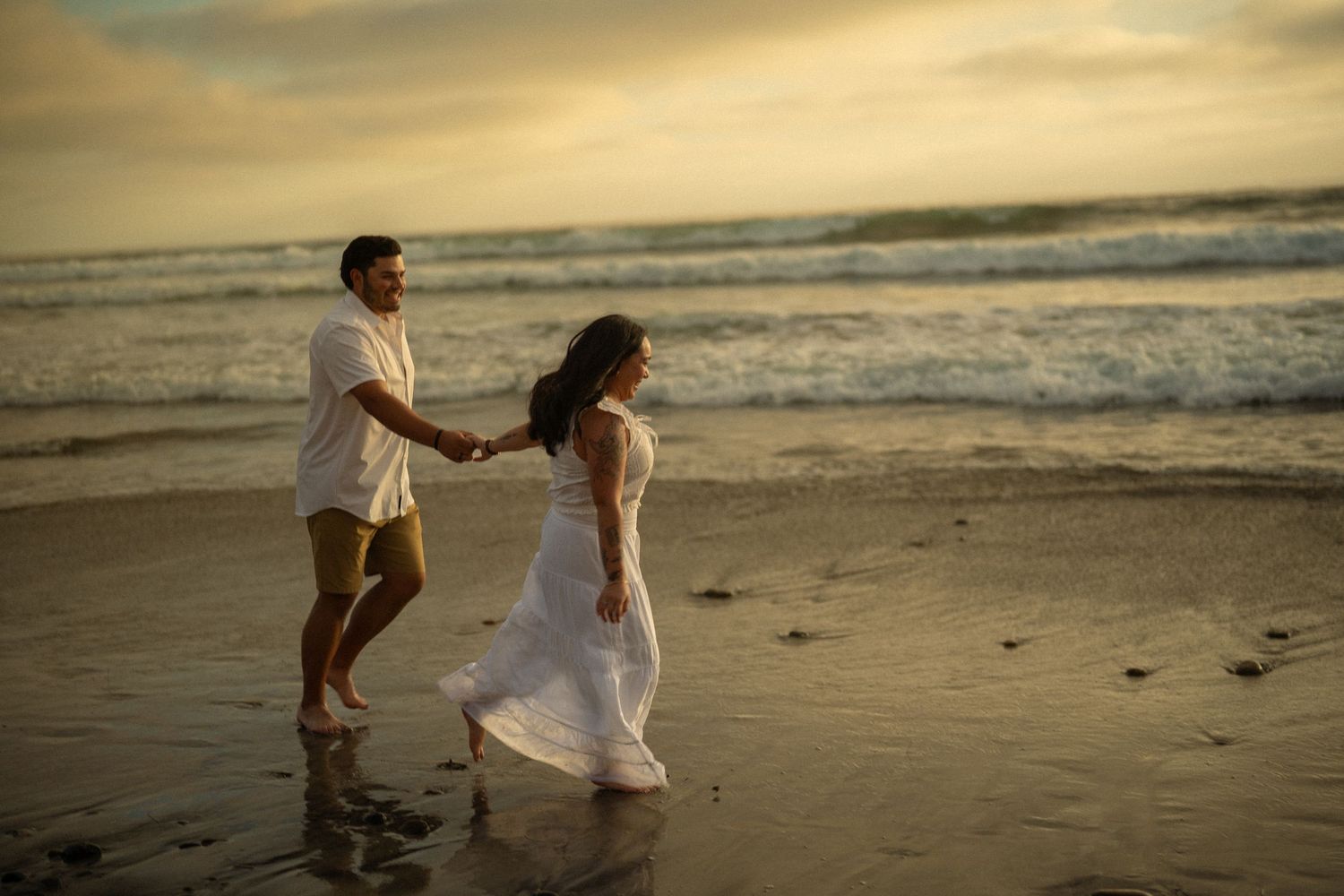 Couple holding hands walking on a beach at sunset, woman in white dress, man in casual attire.