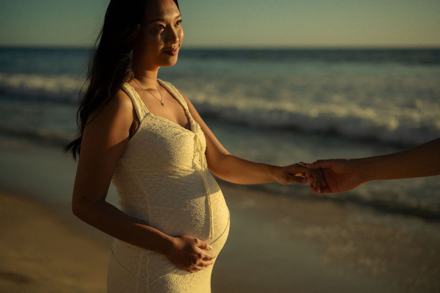 Pregnant woman in white lace dress holding hands on beach at sunset, cradling baby bump near ocean waves.