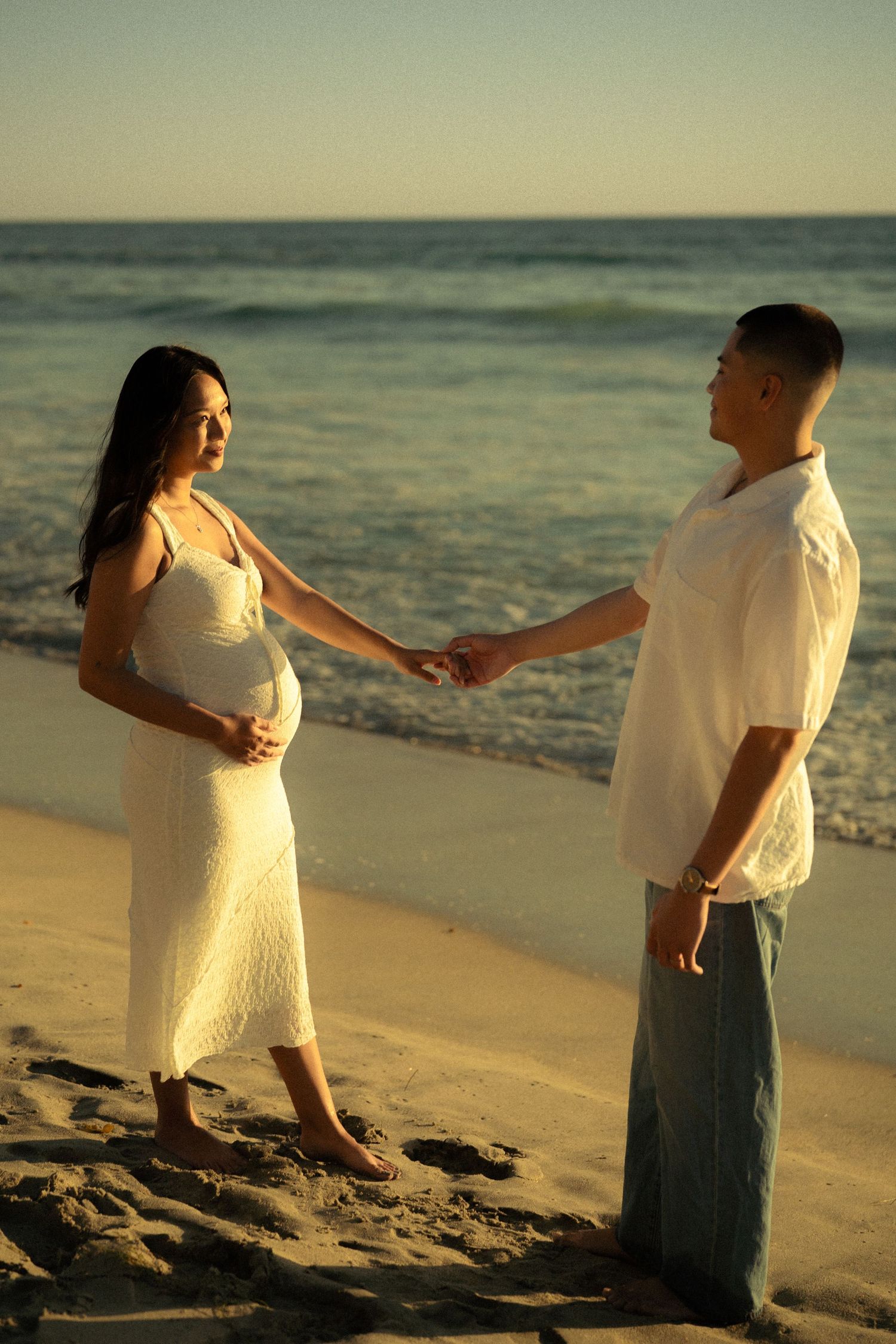 Pregnant woman in white dress holding hands with partner on beach at golden hour sunset.