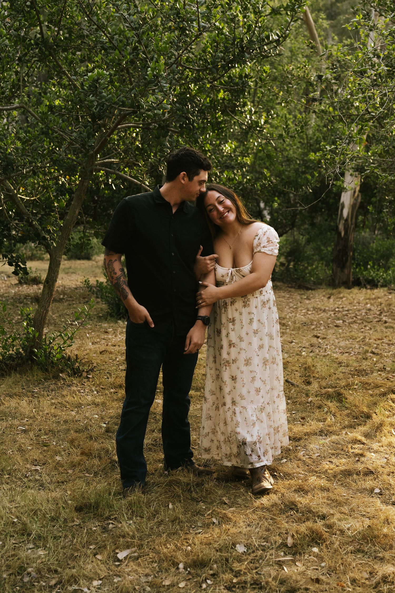 Couple embracing outdoors in a sunny orchard, woman in floral dress laughing as man kisses her cheek.