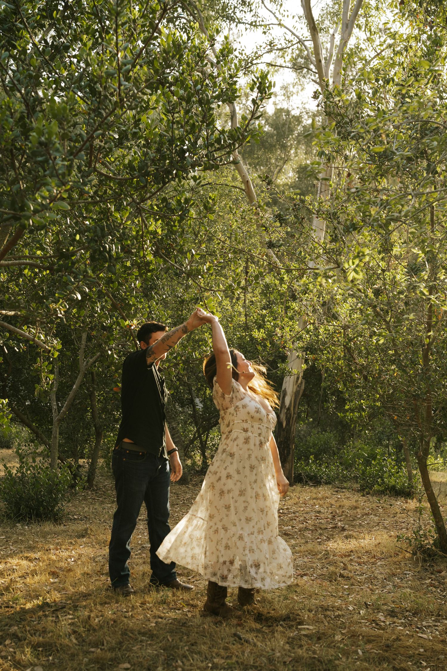Couple dancing together in sunlit forest clearing, man spinning woman in floral dress among green trees.