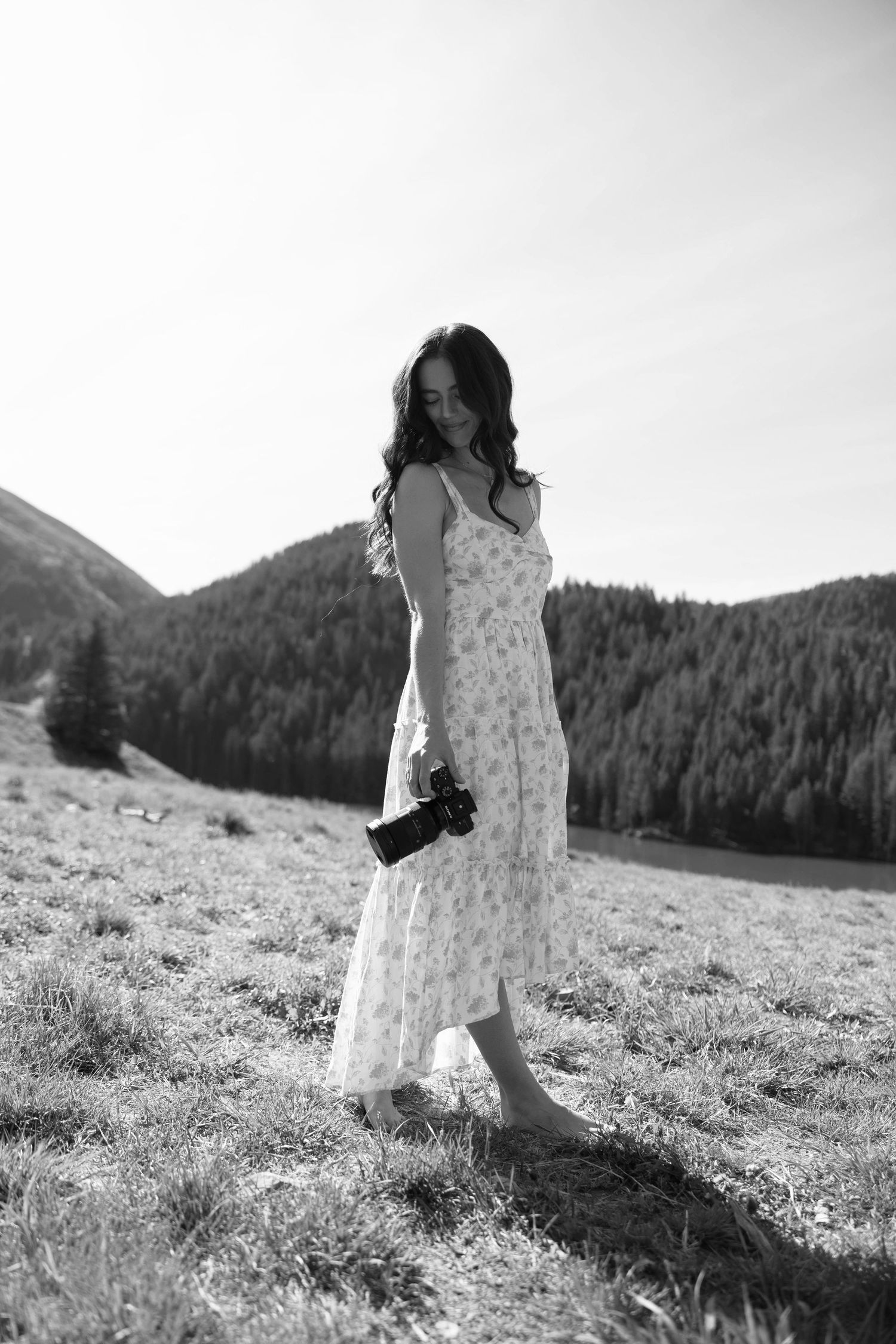 Black and white photo of a young woman in a floral dress holding a camera, standing in a mountain meadow.