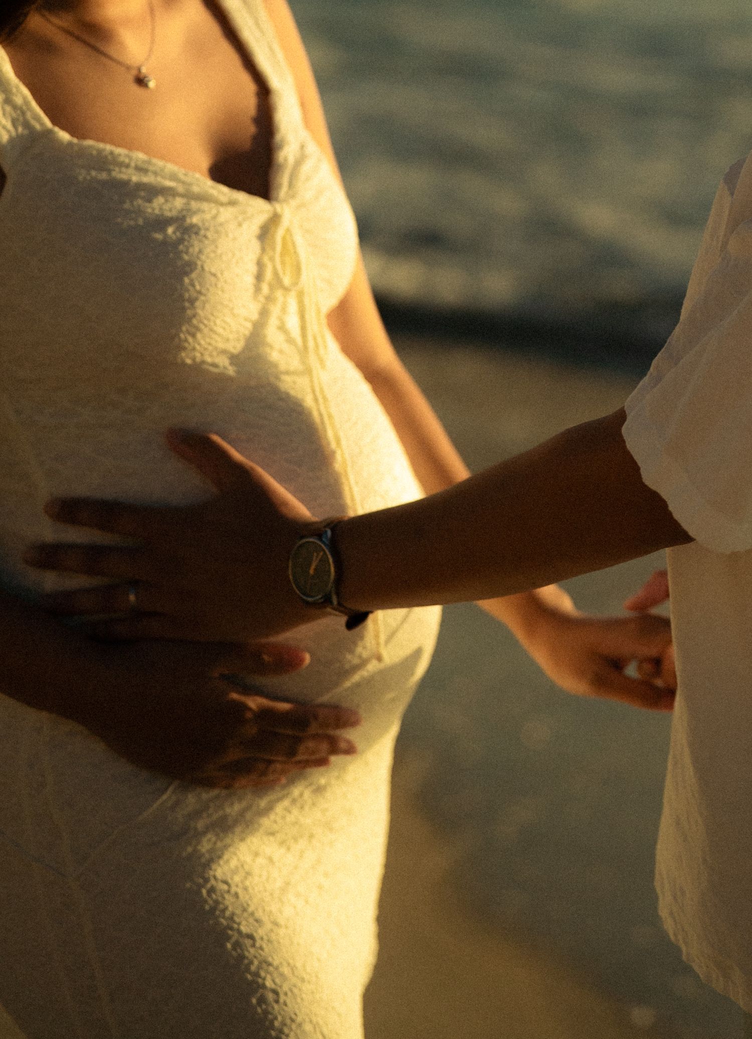Pregnant woman in white dress on beach with partner's hand on baby bump at golden hour.