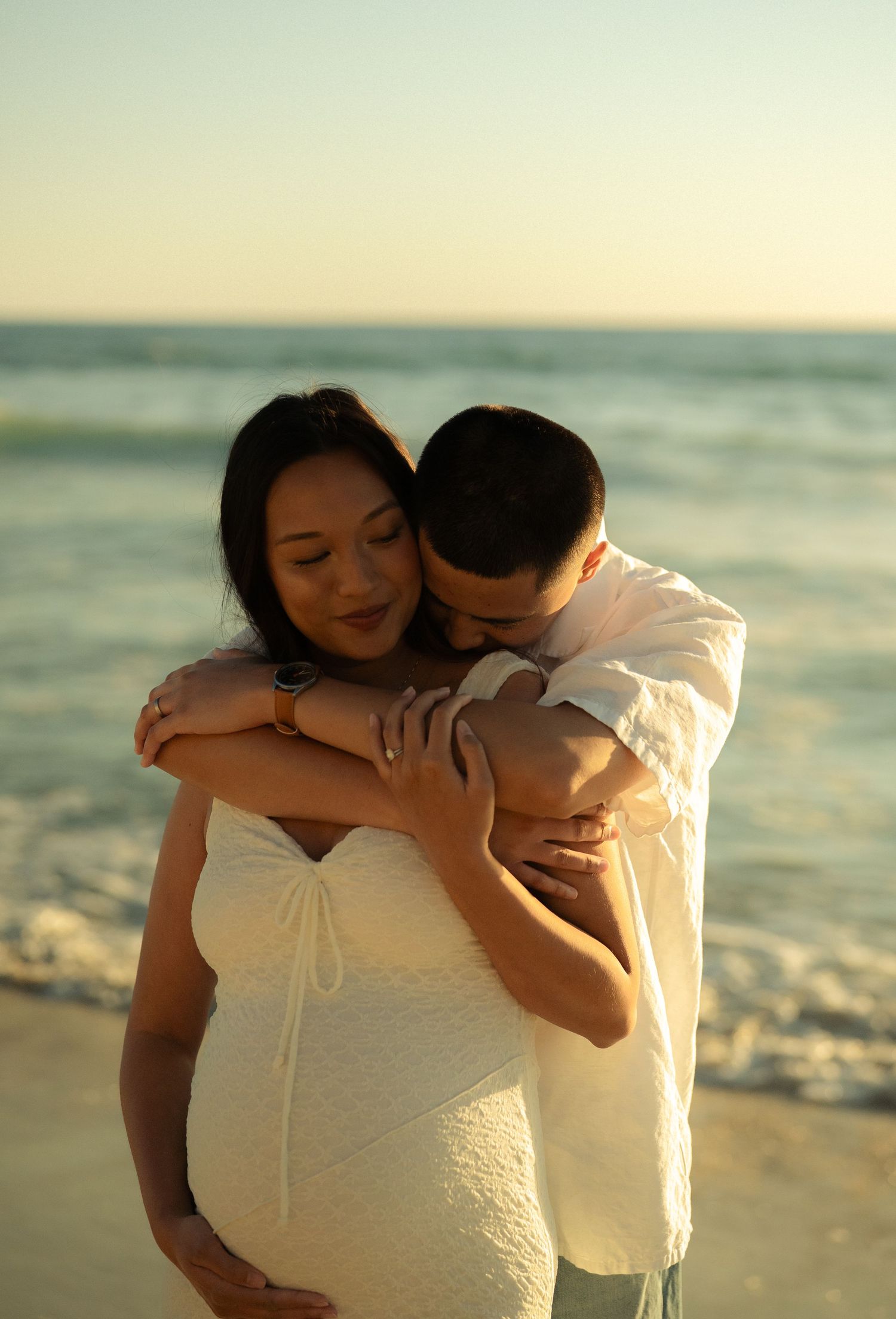 Man tenderly hugging pregnant woman in white dress from behind on beach at golden hour near ocean waves.