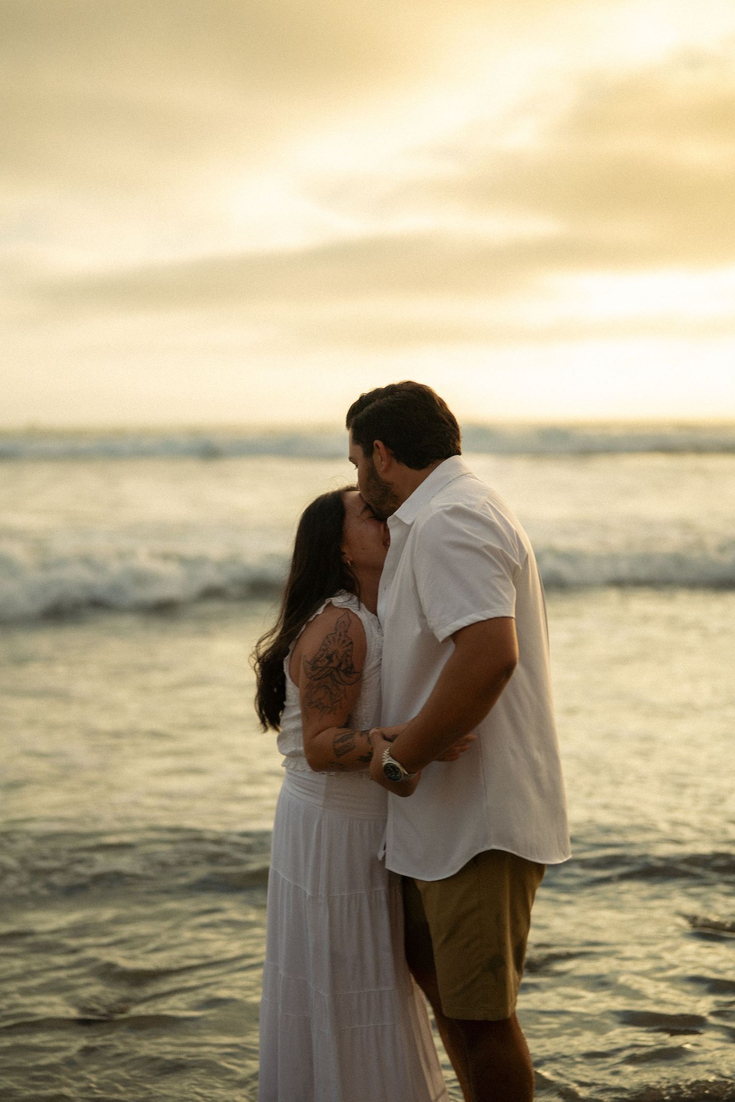 Couple sharing a romantic kiss on the beach at golden sunset, woman in white dress, ocean waves in background.