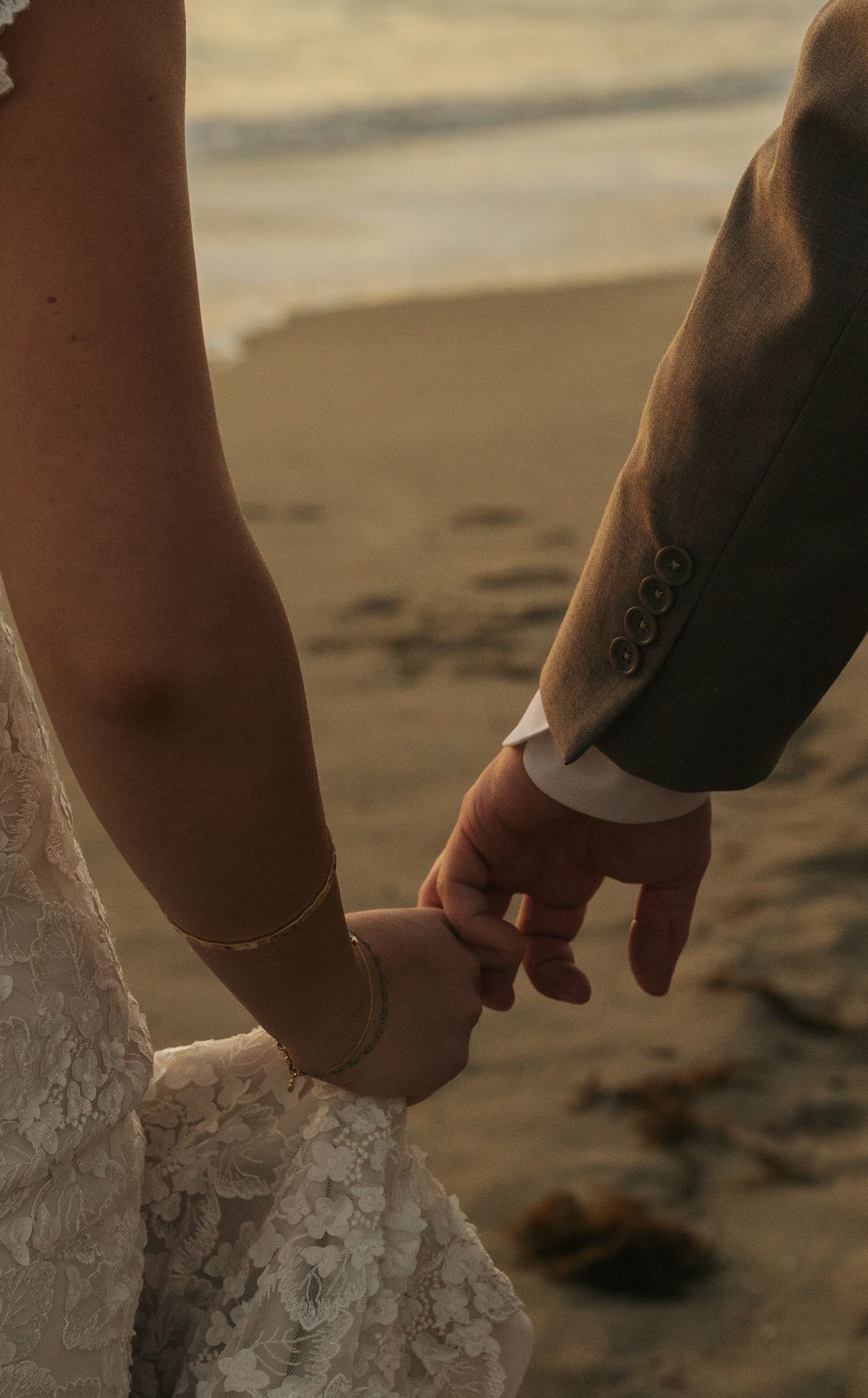 Close-up of bride and groom holding hands on sandy beach, bride wearing delicate gold bracelet and lace dress.