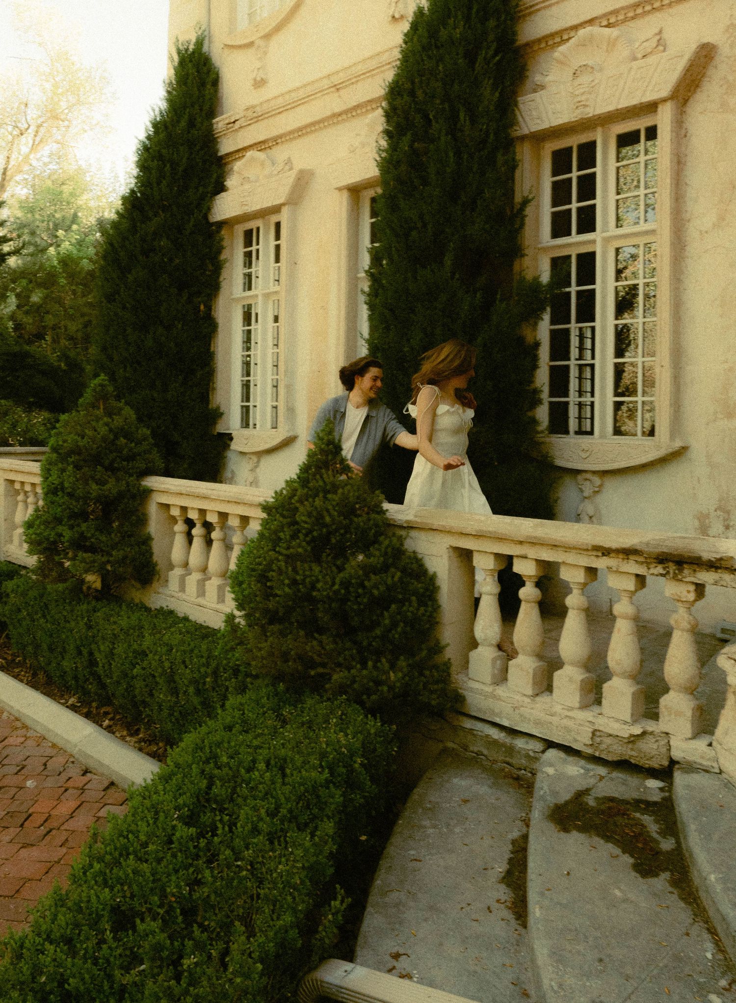 A couple shares a moment on an elegant mansion balcony surrounded by cypress trees.