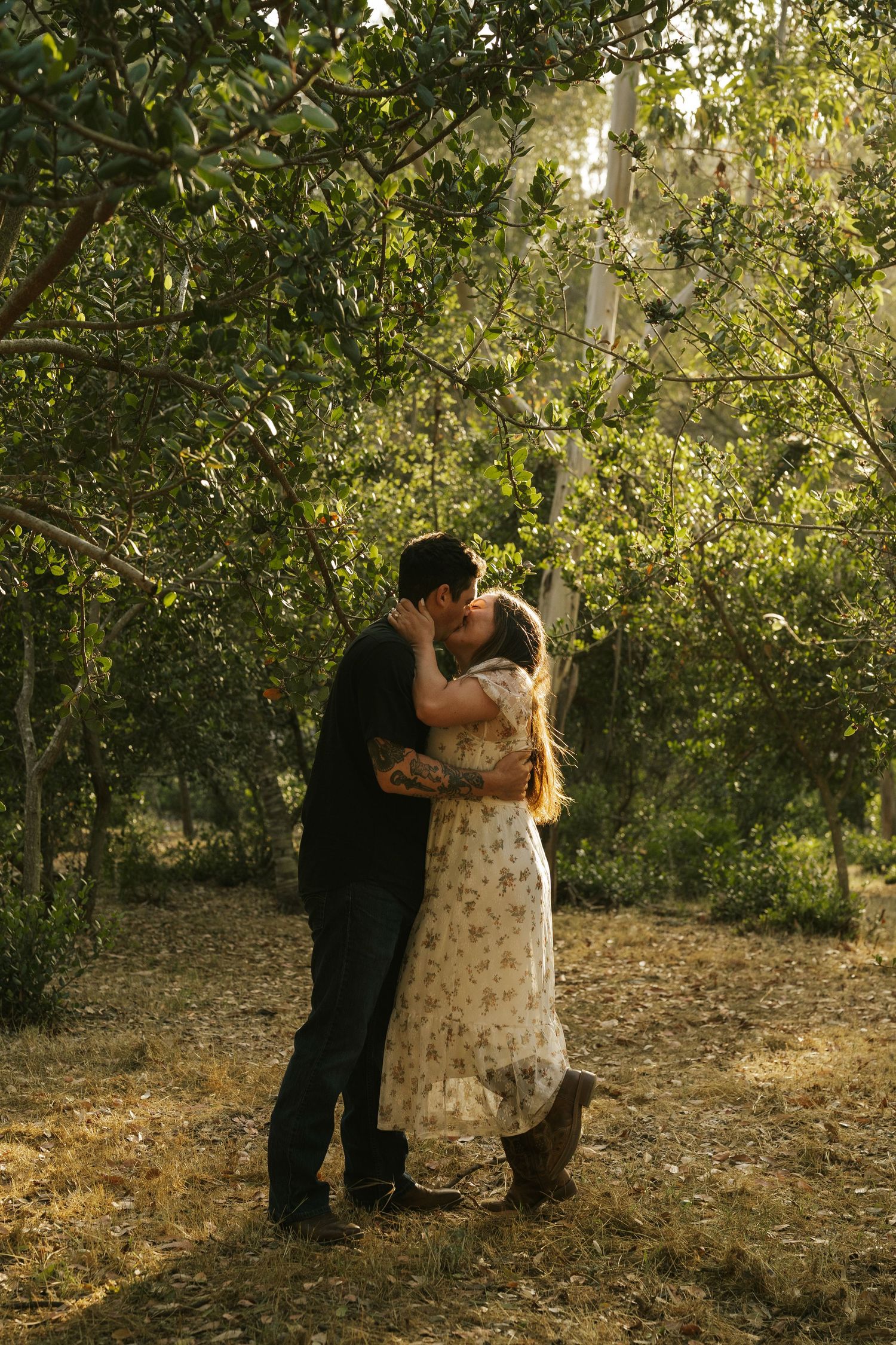 Couple sharing a romantic kiss surrounded by lush green trees in a sunlit outdoor garden setting.