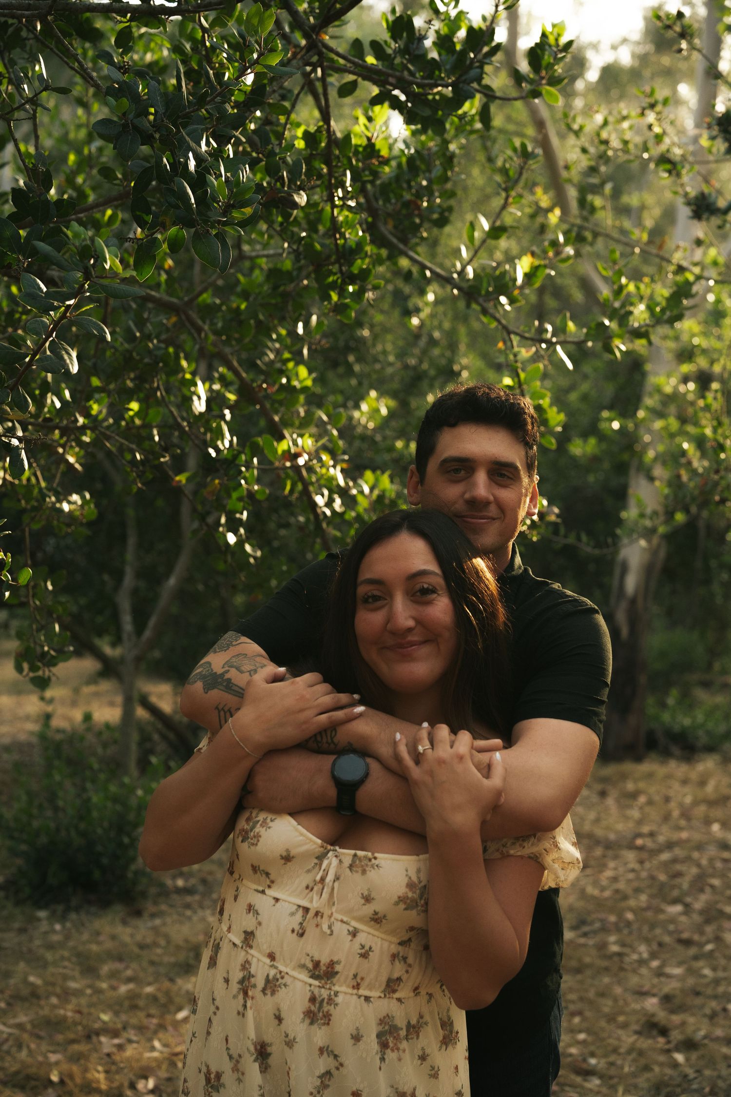 Couple embracing in front of lush green trees, woman in floral dress with tattoo, man in black.
