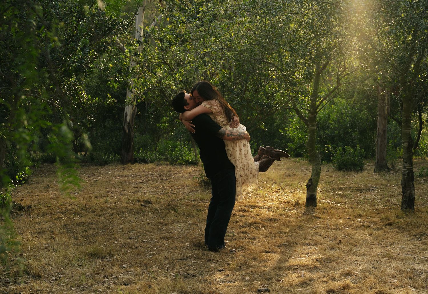 Romantic couple embracing in sunlit forest, man lifting woman among green trees on golden ground.