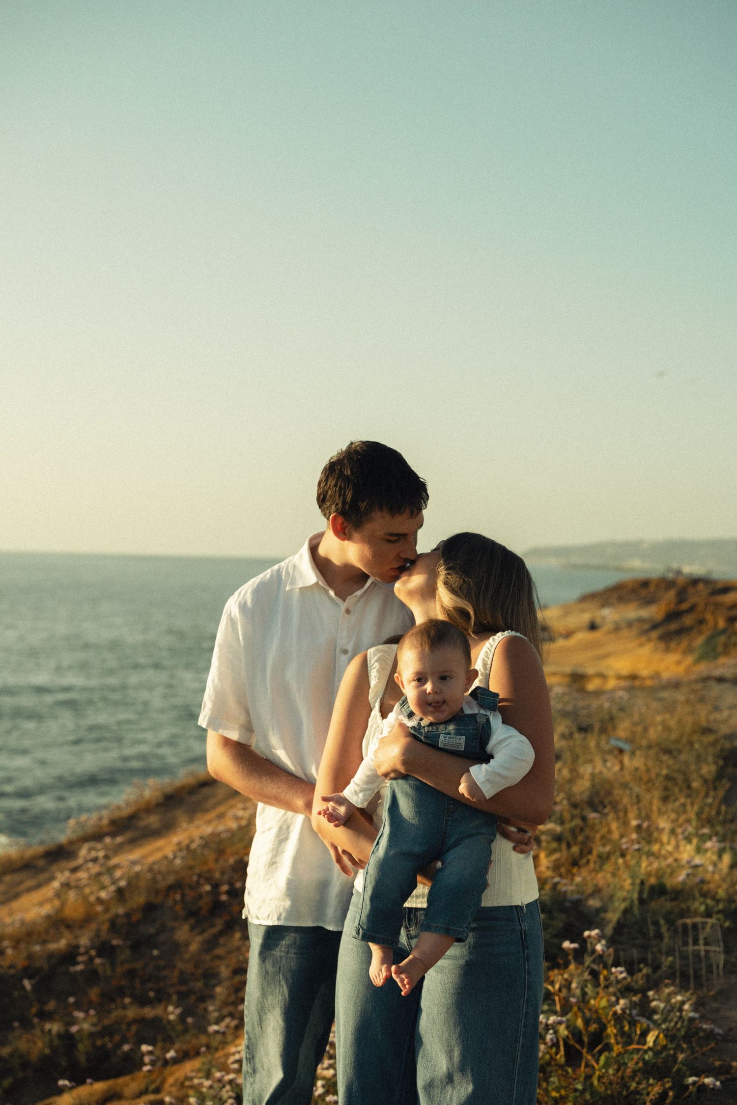 Young couple kissing while holding their baby on a scenic coastal cliff at golden hour.