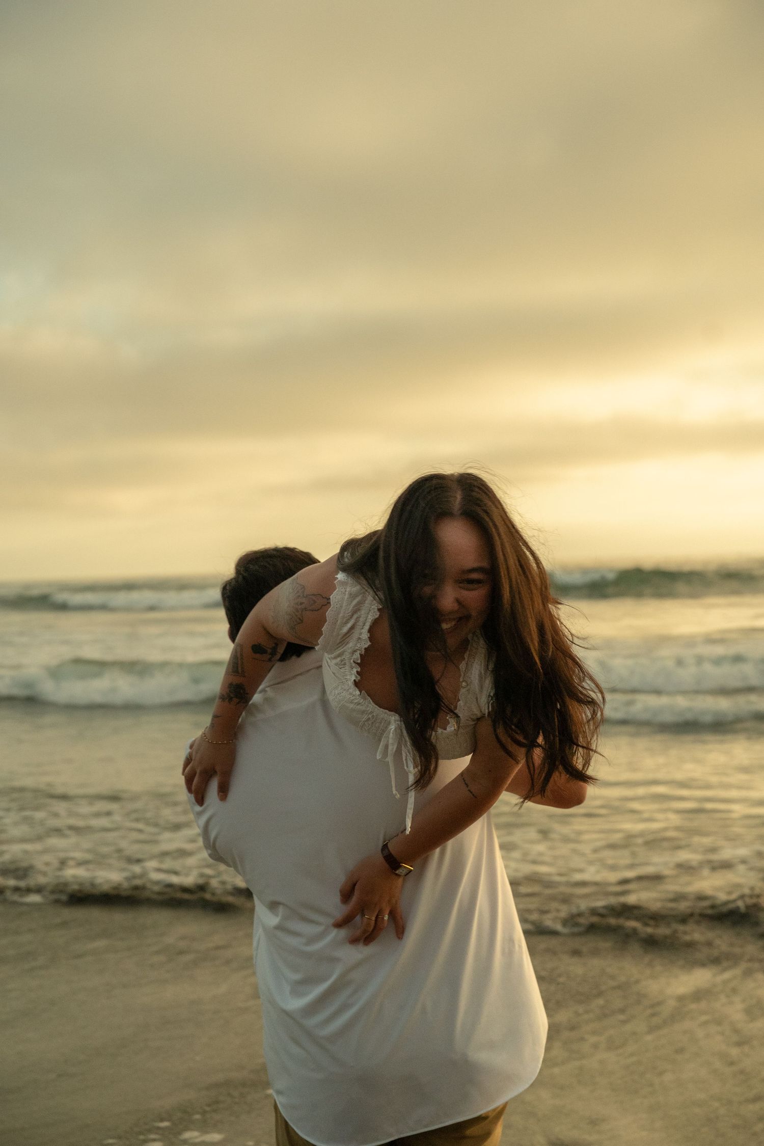 Joyful couple laughing on a beach at sunset, man lifting woman in a white dress by the ocean.