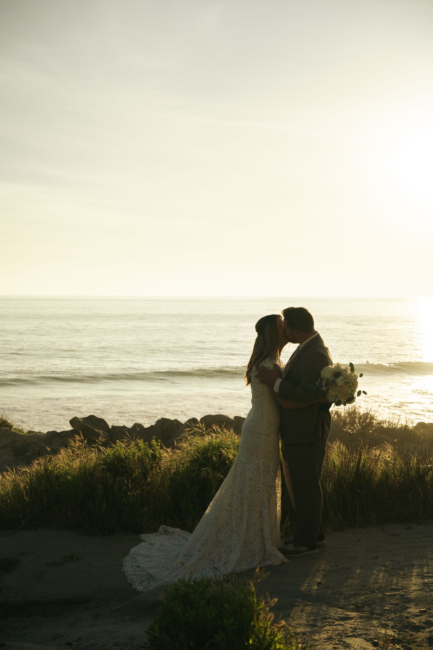 Bride and groom share a romantic sunset kiss on a rocky beach, bride holding a white bouquet.