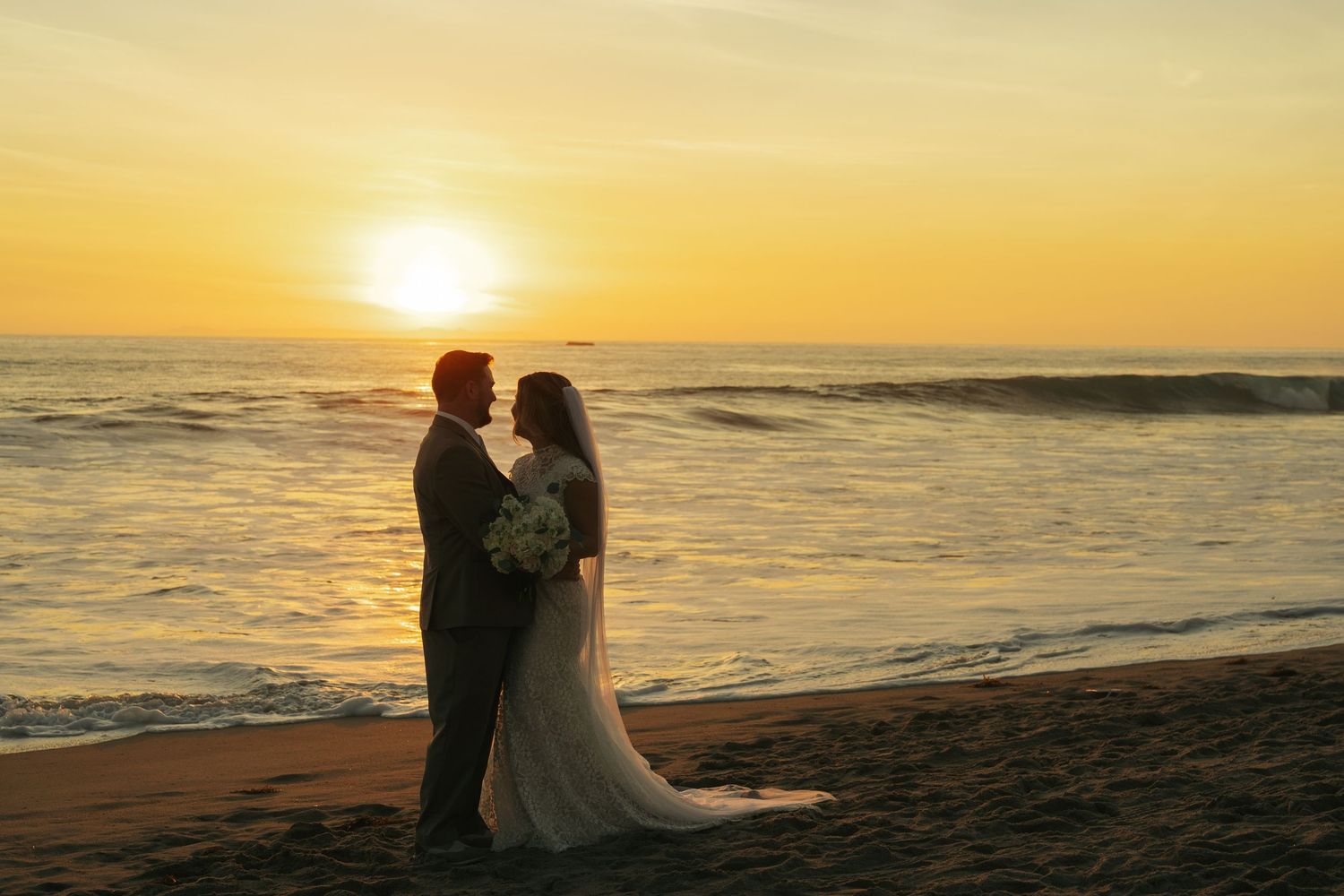Bride and groom silhouetted against a golden sunset on a beach, sharing a romantic moment by the ocean waves.