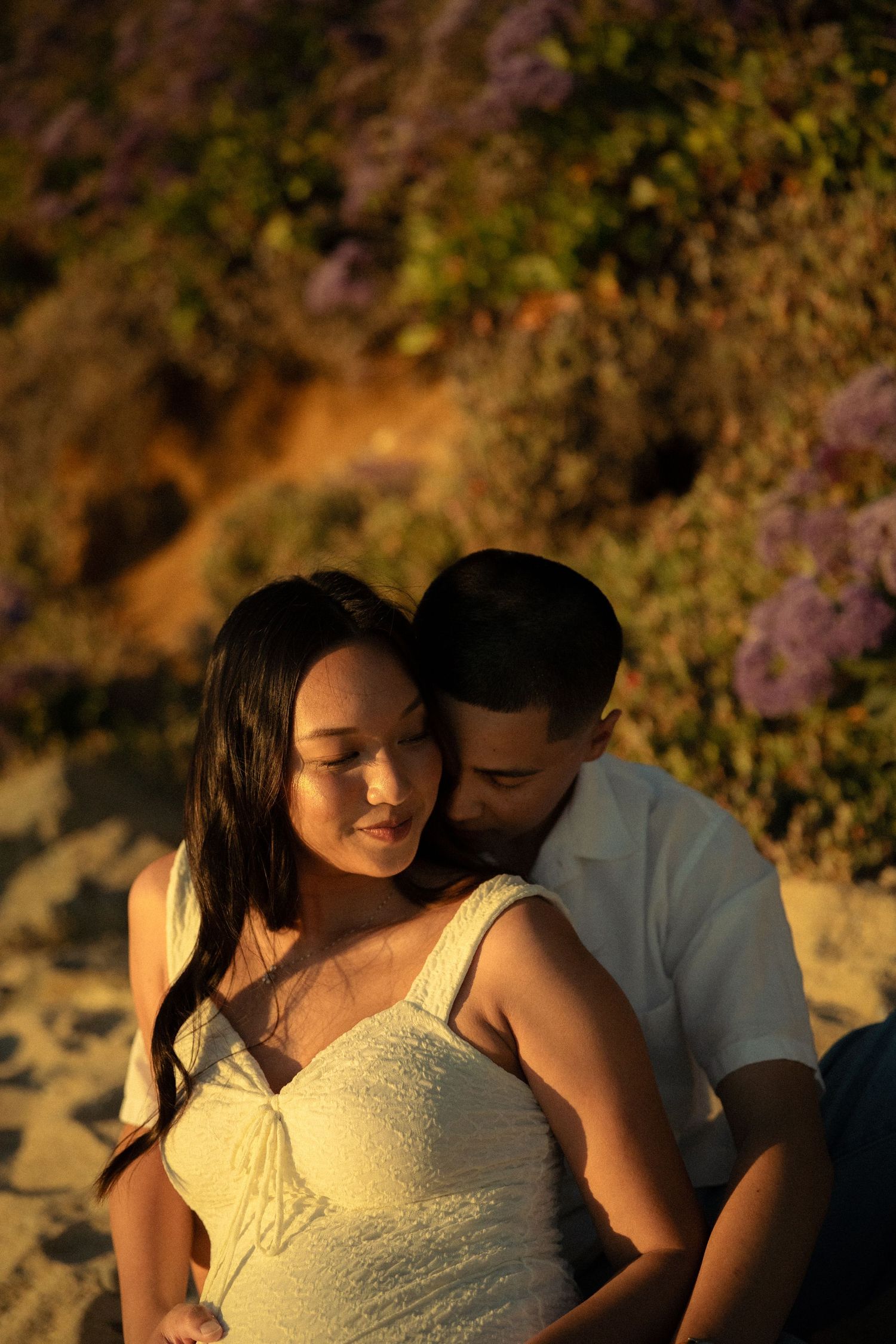 Couple sitting on sandy beach at golden hour, woman in white dress leaning back against man amid purple wildflowers.