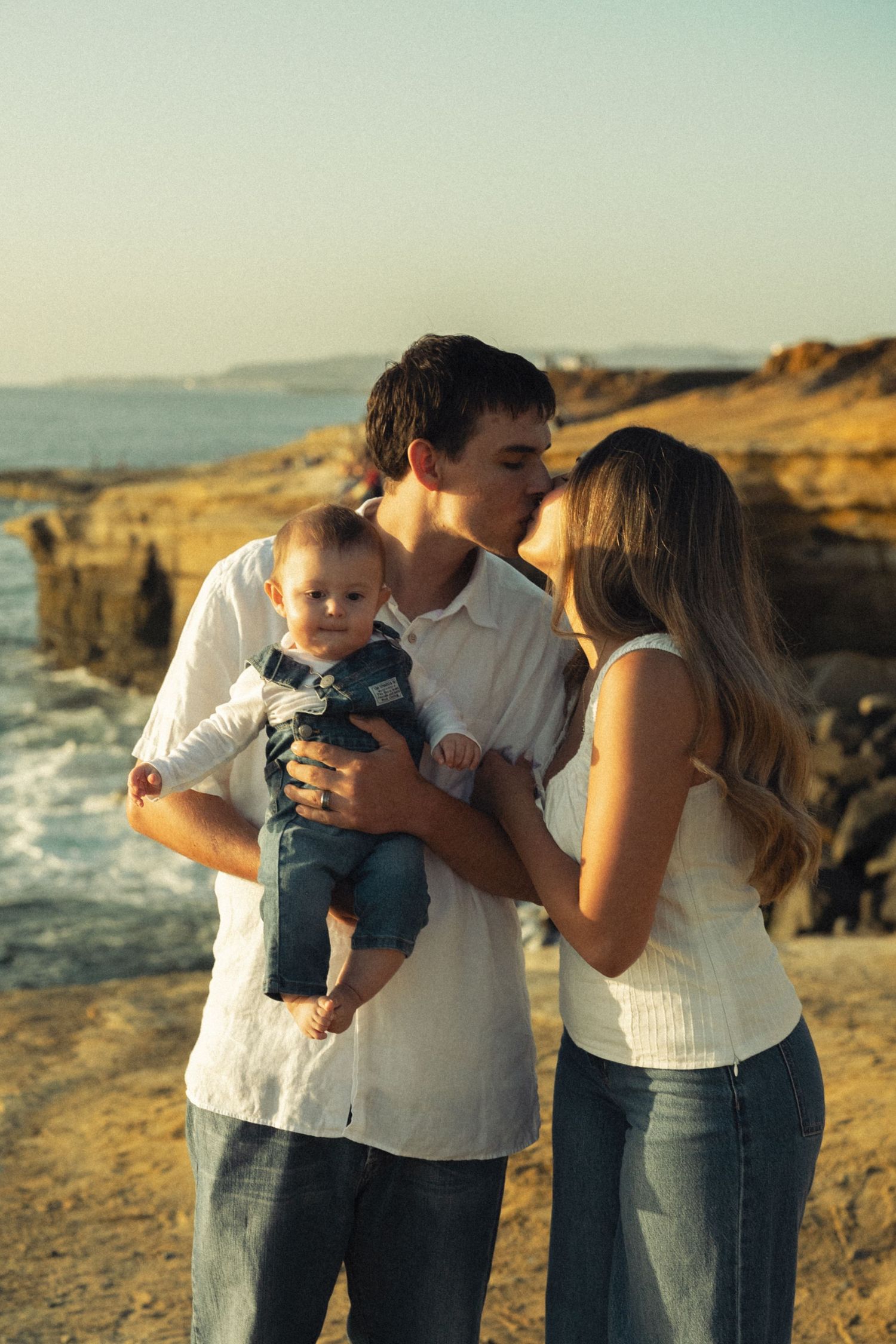 Young parents kissing while father holds their infant baby on a scenic rocky beach at golden hour.