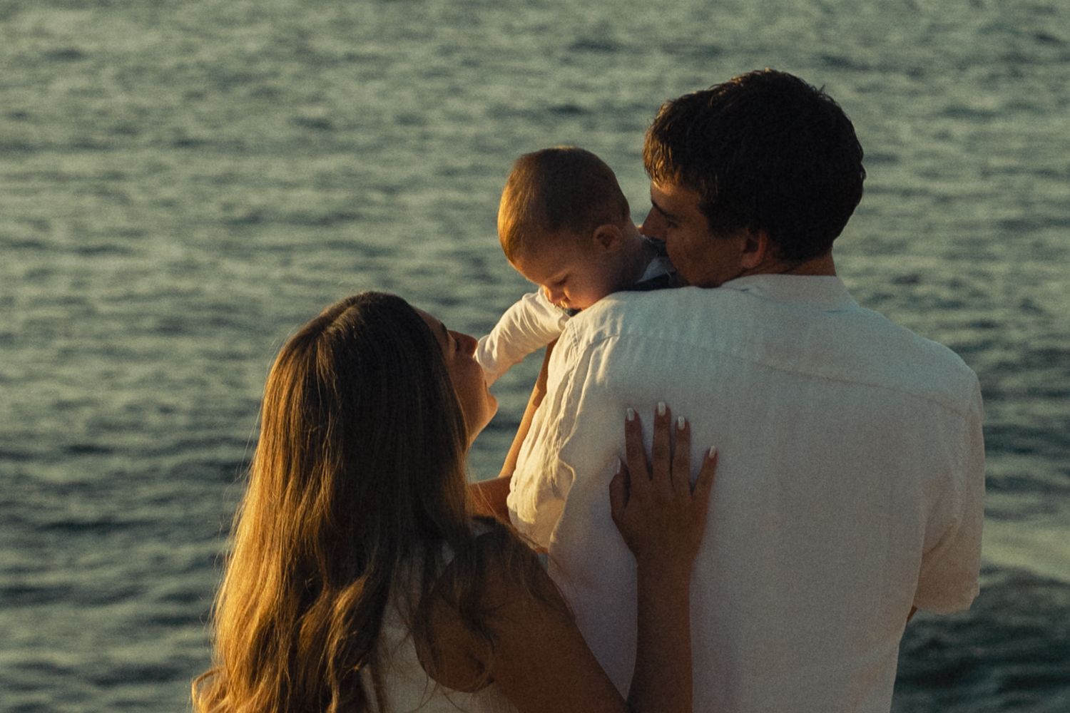 Family with baby enjoying a sunset by the ocean, parents holding infant while gazing at the sea.
