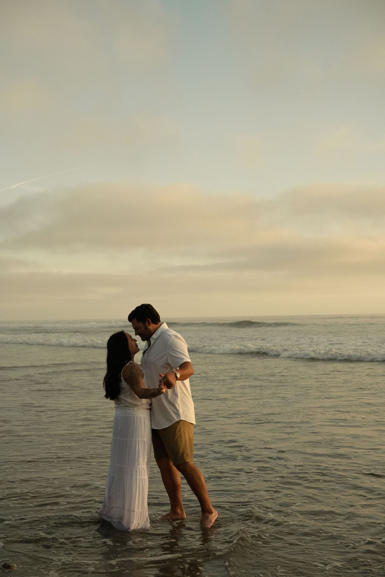 Couple embracing and kissing at the beach shore during golden sunset, woman in white dress, man in white shirt.